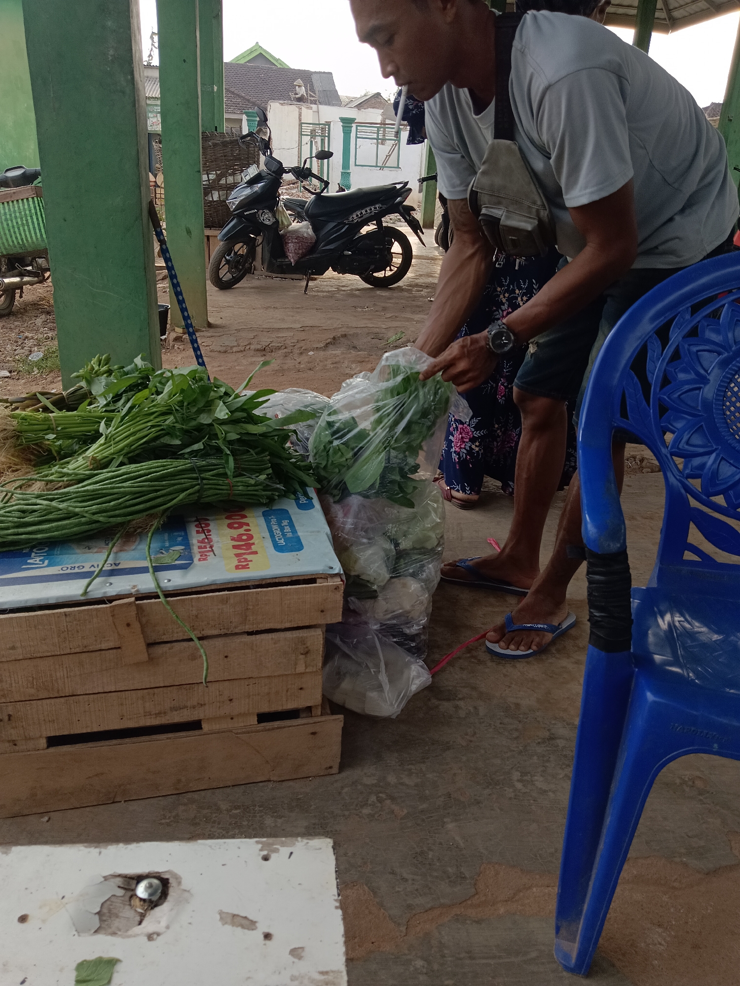 A medium shot captures a man in a light blue t-shirt and dark shorts, standing in what appears to be an outdoor market stall in Taman Cari, Indonesia. He is in the process of bagging produce, holding a clear plastic bag filled with green leafy vegetables in his right hand and reaching for more with his left. A light brown crossbody bag is slung over his shoulder, and a watch is visible on his left wrist. In the background, a black motorcycle is parked, and a wooden crate filled with produce sits in the foreground. A bright blue plastic chair is positioned to the right of the frame. The scene is set against a backdrop of buildings and vegetation, suggesting a typical street market environment.