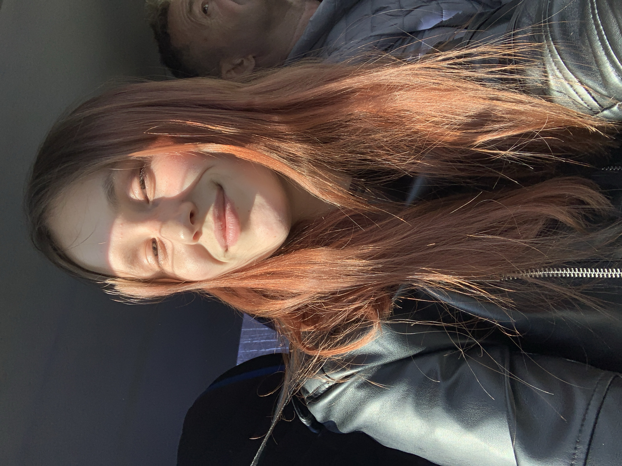 The image features a young woman with long, reddish-brown hair, predominantly lit by bright, warm sunlight. She is smiling gently, with her eyes partially closed due to the intensity of the light. Her face is well-illuminated, highlighting her features, and her hair catches the light with coppery tones. She is wearing a black leather jacket, the texture and a zipper detail visible towards the bottom right of the frame.
In the upper background, an older person's head is partially visible and slightly out of focus. One eye and part of their forehead are discernable, and they appear to be looking slightly upwards.
The setting is an interior space with a dark, featureless background, possibly within a vehicle or a room. The strong, direct illumination suggests it is daytime, likely morning or late afternoon, with sunlight streaming in. The woman's smile conveys a sense of happiness or contentment. There is no visible text in the image.