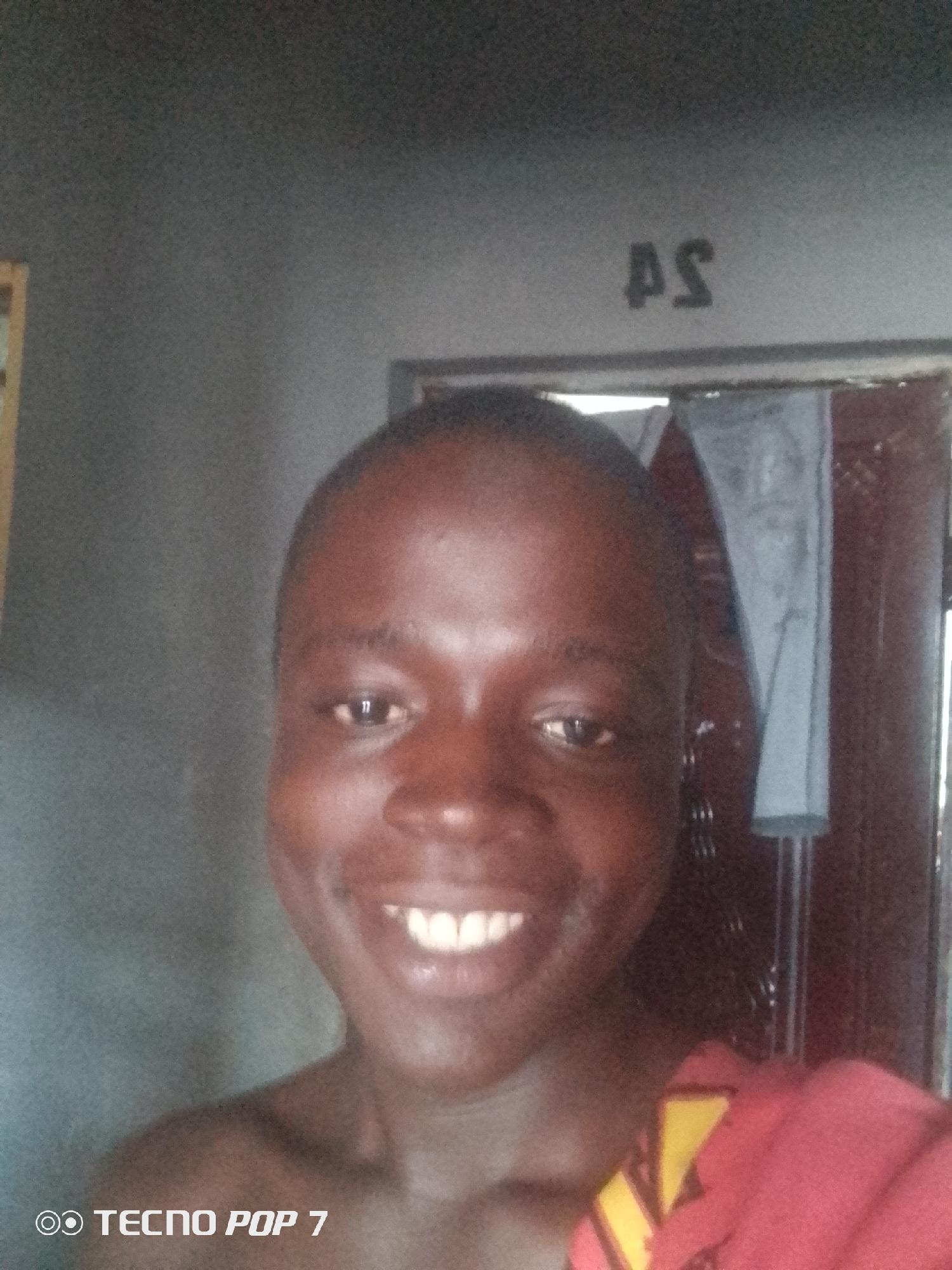 This is a close-up selfie of a young Black person, likely male, smiling directly at the camera. The subject has dark skin, short hair, and is bare-chested, wearing a red garment with yellow accents around their neck and shoulder. Their teeth are clearly visible in their wide smile, and their eyes are dark.
The background appears to be an indoor setting. The wall behind the subject is a neutral gray, with a faint texture. High on the wall, towards the right side, the letters "AS" are visible, written in black. To the right of the subject, a doorway or opening is present, with a dark frame and what looks like a hanging fabric or curtain inside. The lighting suggests it is daytime, with the light source likely coming from the front or slightly to the side, illuminating the subject's face.
In the bottom left corner of the image, the text "TECNO POP 7" is watermarked, indicating the device used to capture the photo. The overall mood is cheerful and relaxed, conveyed by the subject's bright smile. The location context provided is Ipetumodu, Nigeria.