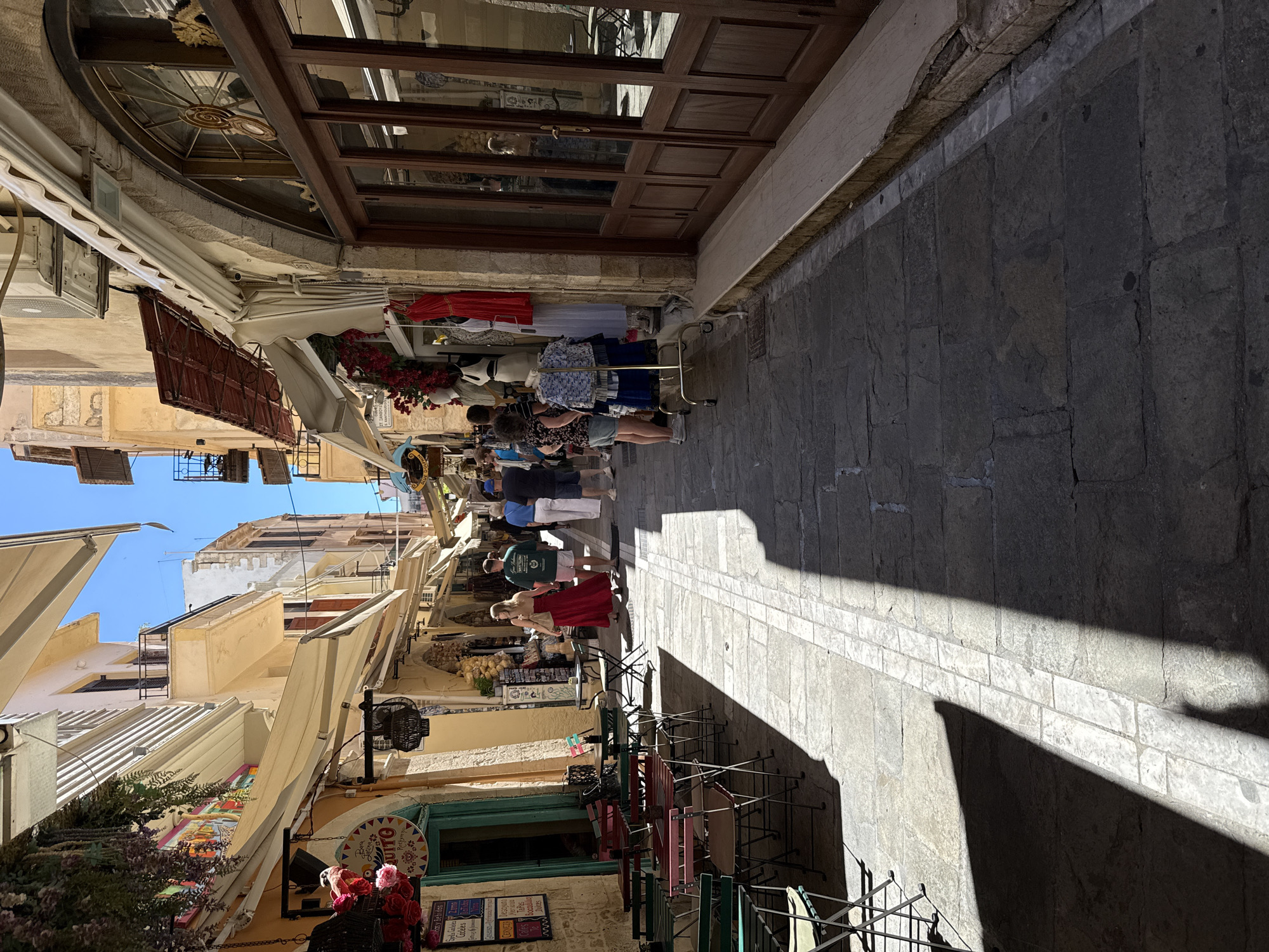 The image shows a sunlit, narrow street in Rethymno, Greece. The street is paved with stone slabs and is lined with shops and cafes on both sides. Many of the buildings have awnings extending over the sidewalks, providing shade. There are outdoor tables and chairs set up in front of a cafe on the left side of the street. On the right side, a shop displays clothing on mannequins and a rack outside. Several people are walking down the street, some are browsing the shops, and others are walking together. The sky is clear and blue, and the sun casts distinct shadows on the pavement. The architecture appears to be a mix of Venetian and Ottoman influences, characteristic of Rethymno's old town.