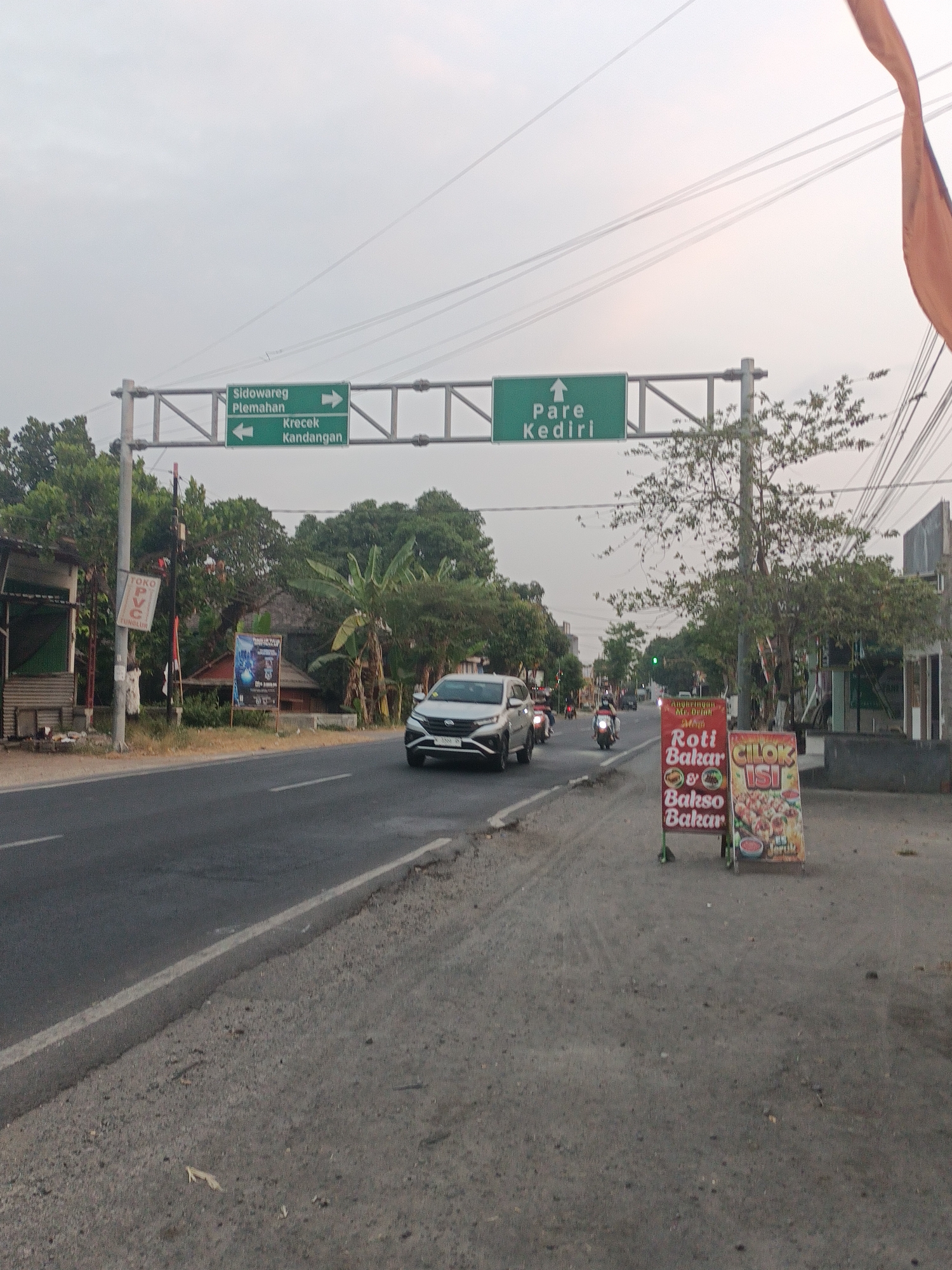 This image shows a street scene in Badas, Indonesia. A directional signpost over the road indicates directions to Sidowareg, Plemahan, Pare, Kediri, Krecek, and Kandangan. To the right of the road, there are two roadside signs advertising food: one for "Roti Bakar & Bakso Bakar" (toasted bread and grilled meatballs) and another for "Cilok Isi" (stuffed meatballs). Several vehicles, including a white SUV and motorcycles, are present on the road. Trees and buildings line the sides of the street, and utility lines are visible overhead. The sky is overcast.