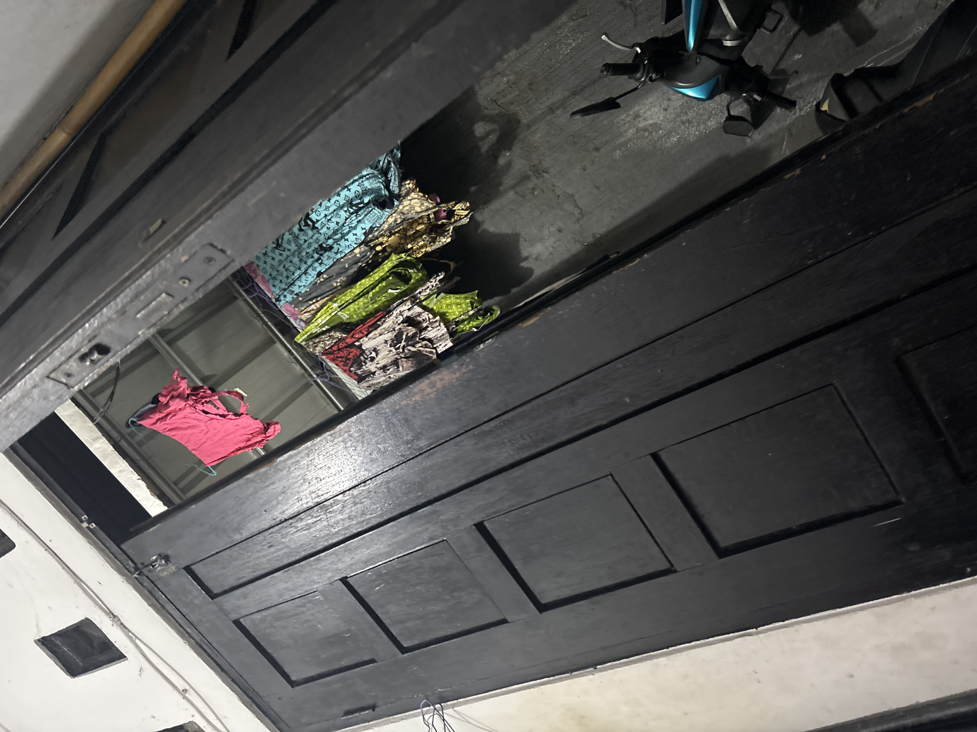 A low-angle, full shot captures a dark wooden door with recessed panels, viewed from a lower vantage point. Above the door, a section of ceiling is visible, revealing a collection of folded fabrics. These fabrics are arranged in a pile, with some appearing to have patterned prints in blue, green, yellow, and red. A single pink t-shirt is hanging on a hanger to the left of the fabrics, suspended by a wire. To the right of the fabrics, a dark motorcycle is partially visible, with its handlebars and seat in focus. The background consists of a concrete floor and a white wall with dark rectangular openings. The scene suggests an indoor space, likely a storage area or an entryway in North Jakarta, Indonesia, with laundry and a motorcycle present.