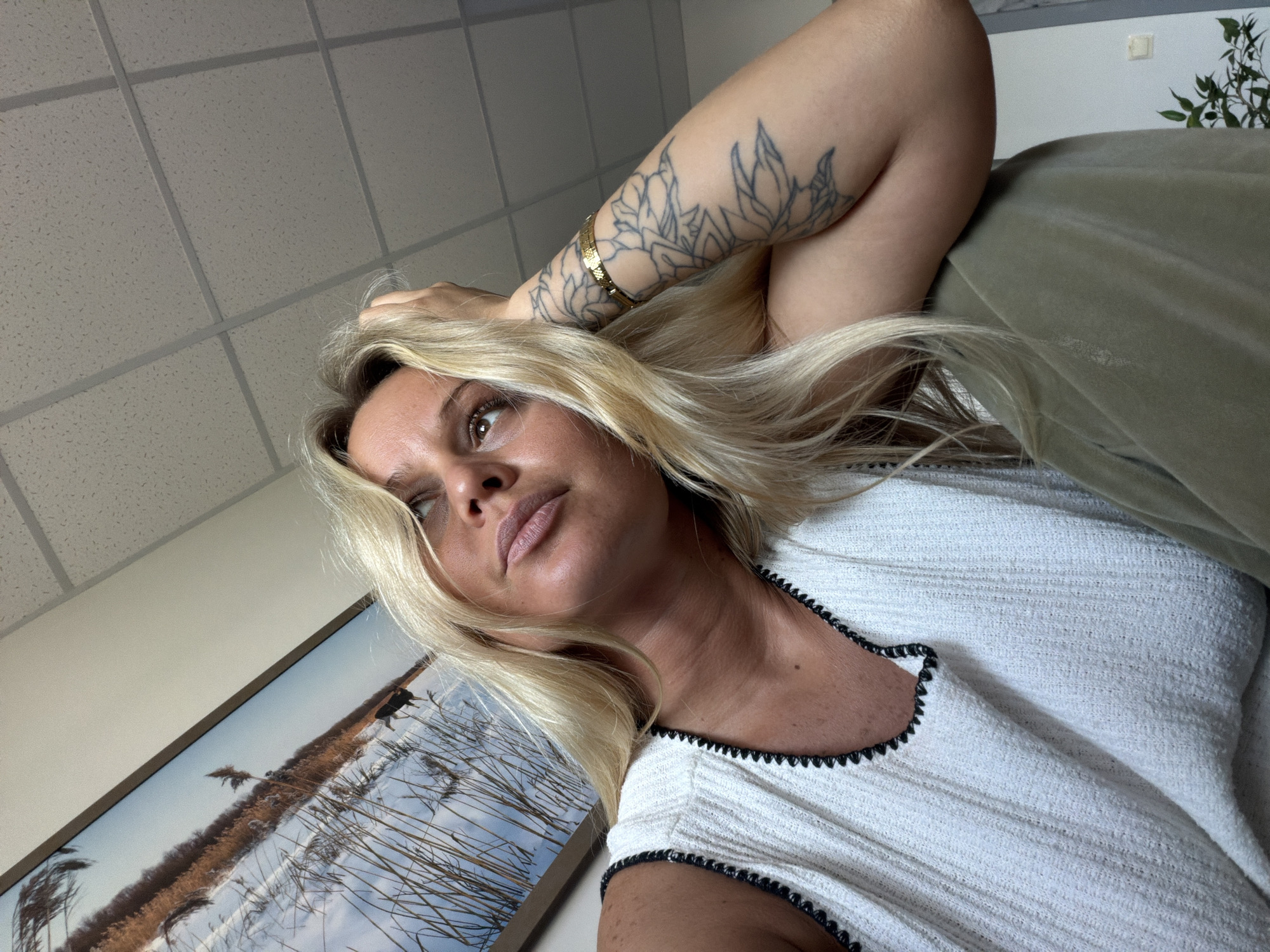 The media file shows a close-up, tilted selfie of a woman with blonde hair, lying down and looking upwards and to her left. She is wearing a white top with black trim. A tattoo of floral designs is visible on her left arm, and she is wearing a gold bracelet on the same arm. The background includes a textured ceiling with square panels and a framed picture on the wall depicting a winter landscape with reeds and snow. A portion of a green sofa or cushion is also visible. The lighting suggests it is daytime. The woman's expression appears thoughtful or relaxed. There is no visible text in the image.