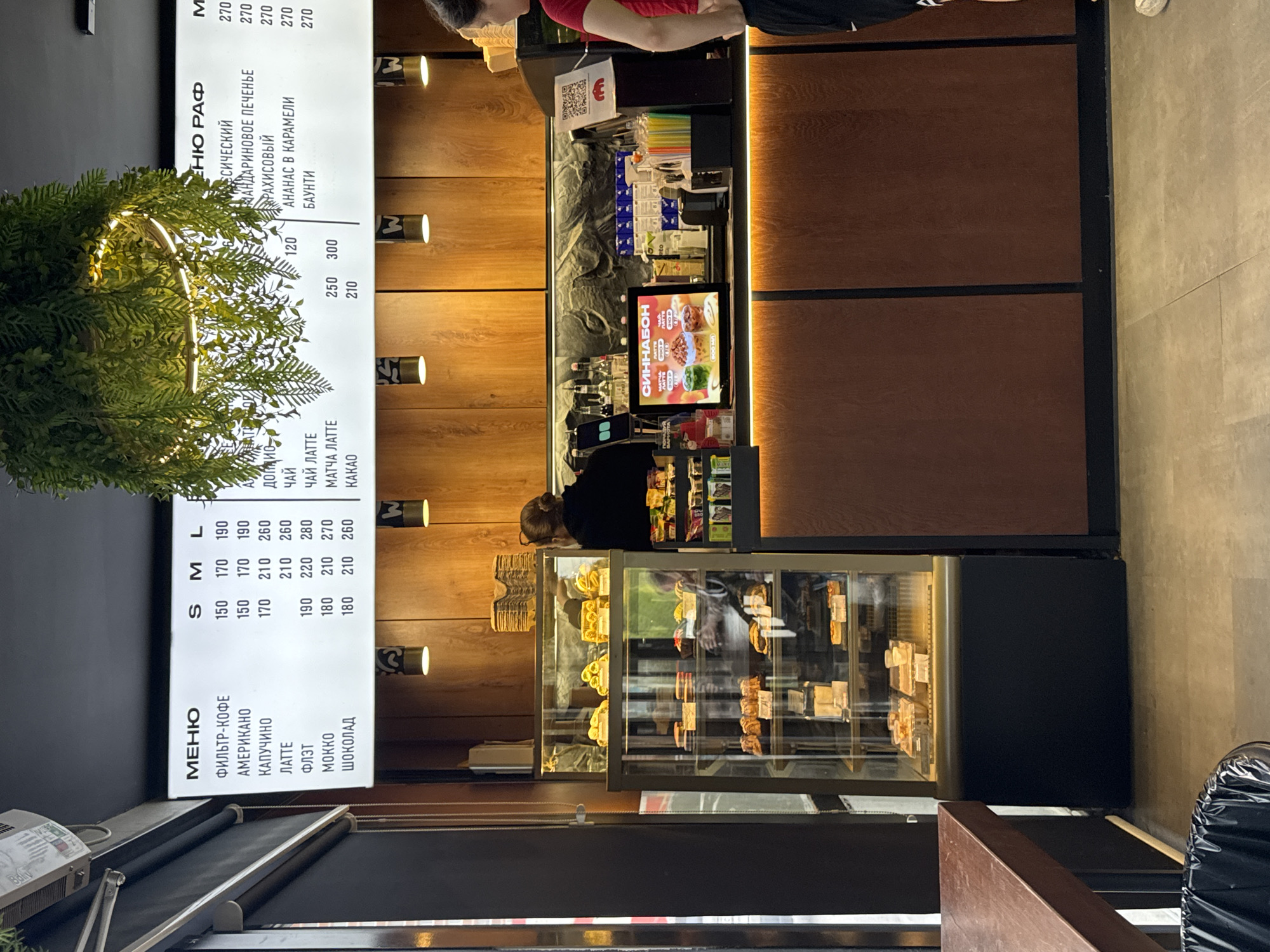 The image captures the interior of a cafe, likely located in Perm, Russia. A menu board displays prices for coffee and other beverages in both small (S), medium (M), and large (L) sizes. The beverages listed include "ФИЛЬТР-КОФЕ" (filter coffee), "АМЕРИКАНО" (Americano), "КАПУЧИНО" (cappuccino), "ЛАТТЕ" (latte), "ФЛЭТ" (flat white), "МОККО" (mocha), and "ШОКОЛАД" (chocolate). A secondary menu board lists different types of "РАФ" (a coffee drink) with names like "ССИЧЕСКИЙ" (classic), "МАНДАРИНОВОЕ ПЕЧЕНЬЕ" (tangerine cookie), "РАХИСОВЫЙ," "АНАНАС В КАРАМЕЛИ" (pineapple in caramel), and "БАУНТИ" (Bounty).
Behind the counter, an employee in a black shirt appears to be working. A screen displays an advertisement for "СИННАБОН" (Cinnabon). The counter includes items such as straws, and a display case contains a variety of pastries. In the foreground, there is greenery in a pot.