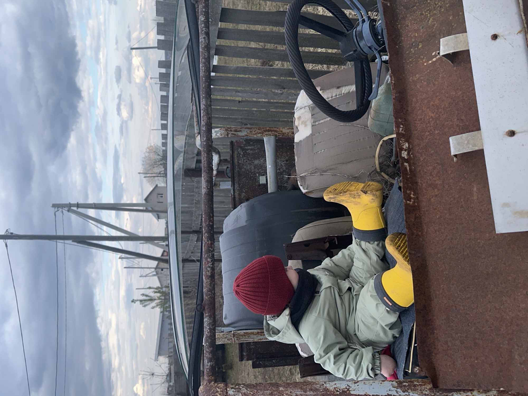 A child in a red knit cap, light green jacket, and yellow rubber boots sits in an old, rusty tractor. The tractor has a black steering wheel, a faded white seat, and a large black tank behind the child. The background shows a cloudy sky, utility poles, and buildings typical of a rural setting. The location is Ludorvay, Russia.