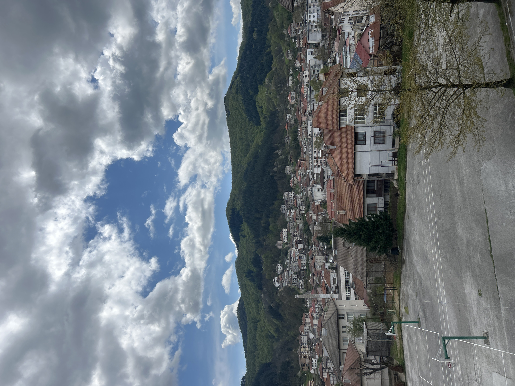 The image shows a view of Zlatograd, Bulgaria, featuring a mountain range covered in green foliage that spans the center and left of the frame. Above the mountain, there is a blue sky with scattered white clouds. A dense collection of white buildings with orange and red tile roofs extends from the mountain's base across the middle and right side of the picture. A basketball court is visible in the lower right corner, with visible lines and two green basketball hoops. Bare trees are present alongside the court. The image appears to be taken from an elevated vantage point, possibly from a building overlooking the town and mountain.