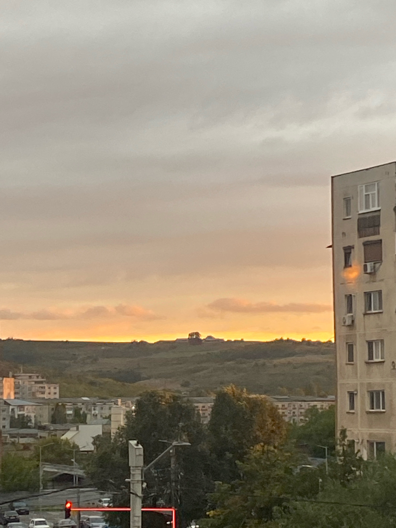 The image shows a wide view of a city with a dramatic sunset in the background, likely taken from a higher vantage point overlooking the city of Iași, Romania. The sky is filled with muted, warm tones of orange and yellow, with soft clouds. In the foreground, a tall, weathered apartment building dominates the right side of the frame, its windows reflecting the sunset light. Below the apartment building, a dense cluster of trees obscures the lower levels of other residential buildings. In the mid-ground, a hilly landscape dotted with more apartment buildings stretches across the horizon. The lower portion of the image shows a street level view with cars, a traffic light showing red, and a traffic camera. The overall impression is of an urban environment transitioning into twilight under a colorful sky.