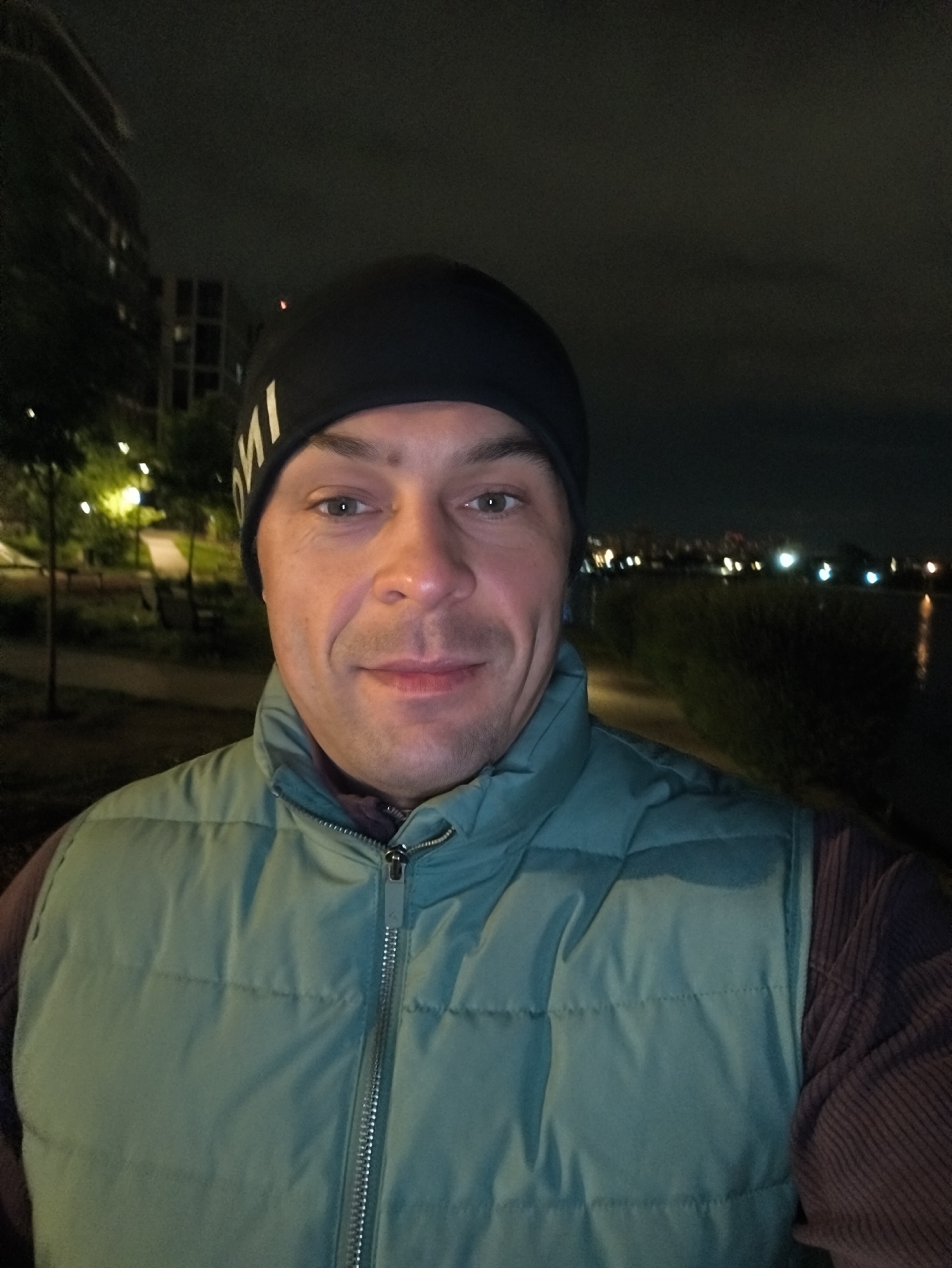 A man is taking a selfie outdoors at night. He is wearing a black beanie, a green puffer vest, and a long-sleeved shirt. Behind him, there are buildings and lights, suggesting a cityscape. Based on the location indicated by the prompt, this scene is potentially in Moscow, Russia. There is also what appears to be a body of water and vegetation in the background.