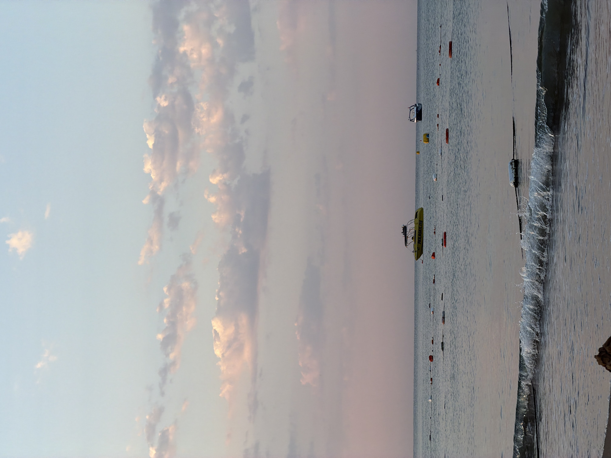 This is a vertical, eye-level shot of the sea in Manavgat, Turkey, during what appears to be sunset or sunrise. The sky is a soft gradient of pale blue to pink, filled with scattered clouds tinged with pink and white. The sea is calm with small waves lapping at the shore in the foreground. Several buoys, some red and orange, and a few white ones, are visible in the water, marking a boundary. Two boats are present: a yellow boat with the word "PARASAILING" visible on its side, located towards the left of the frame, and a smaller white boat further to the right. The water has a grey-blue hue, reflecting the sky. The beach is only visible in the very bottom of the frame, with a small mound of sand at the lower left corner. No people are visible in the scene.