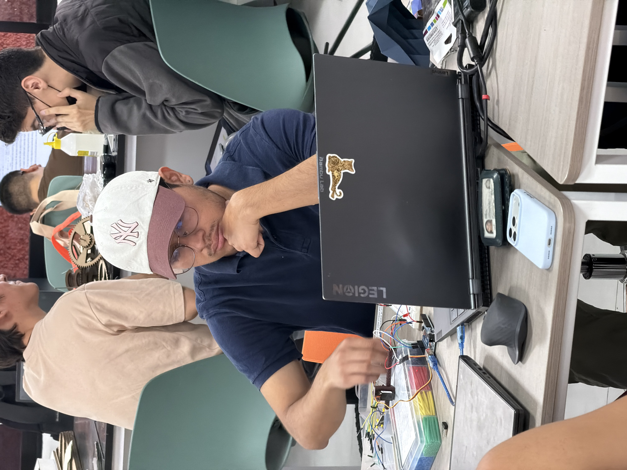 A medium, eye-level shot shows four men around a table inside a building. A man wearing a navy blue polo shirt and a white baseball cap with a burgundy bill works on an electronics project on a breadboard connected to a laptop. A gray wireless mouse and a blue smartphone are on the table next to the laptop. Another man in a tan t-shirt sits to the left of the man working on the project. Behind the man in blue sits a man in a gray jacket with his head bent down. A fourth man is visible in the background to the left, wearing a tan shirt. All four men seem to be focused on the electronics project. The location is assumed to be San Andrés Cholula, Mexico.