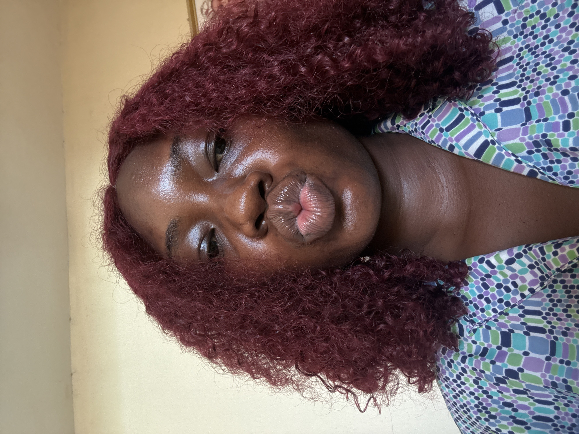 A close-up selfie of a Black woman with reddish-brown curly hair. She has dark skin and full, glossy lips pursed into a kiss. Her eyes are visible, and she appears to be looking directly at the camera. She is wearing a collared shirt with a geometric pattern in shades of blue, green, and purple. The background is a light beige wall. The image suggests an indoor setting, likely a private residence. There is no information or indication that the image was taken in Abuja, Nigeria.