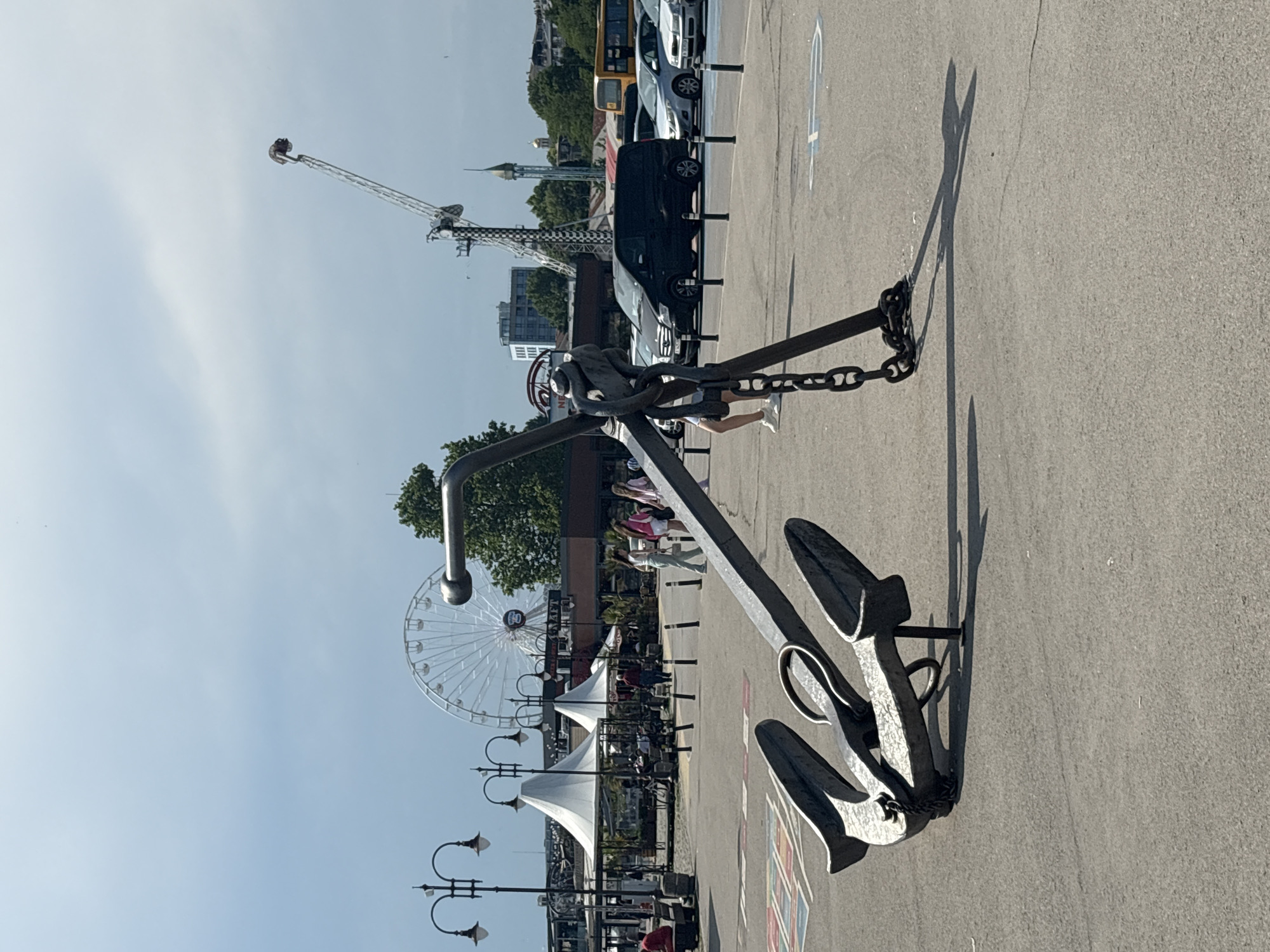 The image captures an outdoor scene on a bright, partly cloudy day in what appears to be a public paved area, consistent with a waterfront or promenade location in a city like Varna, Bulgaria.
In the immediate foreground, a large, dark gray metallic anchor sculpture is prominently displayed, resting on its side. Its stock is oriented vertically, and one of its flukes is firmly on the ground while the other points upwards. A thick, dark metal chain is wrapped around the anchor's shank, extending onto the gray asphalt or concrete pavement where it forms a coil. The anchor and chain cast elongated shadows to the right. Faint painted markings, including what appears to be a rainbow design on the far left and a blue handicapped parking symbol on the far right, are visible on the pavement.
The midground features a bustling pedestrian area. On the left, two ornate, dark-colored street lamp posts, each with multiple luminaires, illuminate the space. Further back, two distinctive white structures with conical roofs, resembling large tents or umbrellas, are situated over what appears to be outdoor seating areas. Behind these, various individuals are walking, some in small groups, and indistinct buildings form the backdrop.
In the right-midground, a parking area is delineated by a line of short, black bollards. Several vehicles are parked, most notably a black minivan or passenger van. A yellow bus is visible further in the background on the far right.
The background is dominated by amusement park attractions. A large, white Ferris wheel stands prominently in the center. To its right, a tall, slender amusement ride extends upwards, featuring a long, angled support structure with a checkered pattern and a capsule at its apex. A smaller, ornate building with a golden dome is visible further to the right. Scattered green trees punctuate the urban landscape. The sky above is light blue with scattered white clouds.