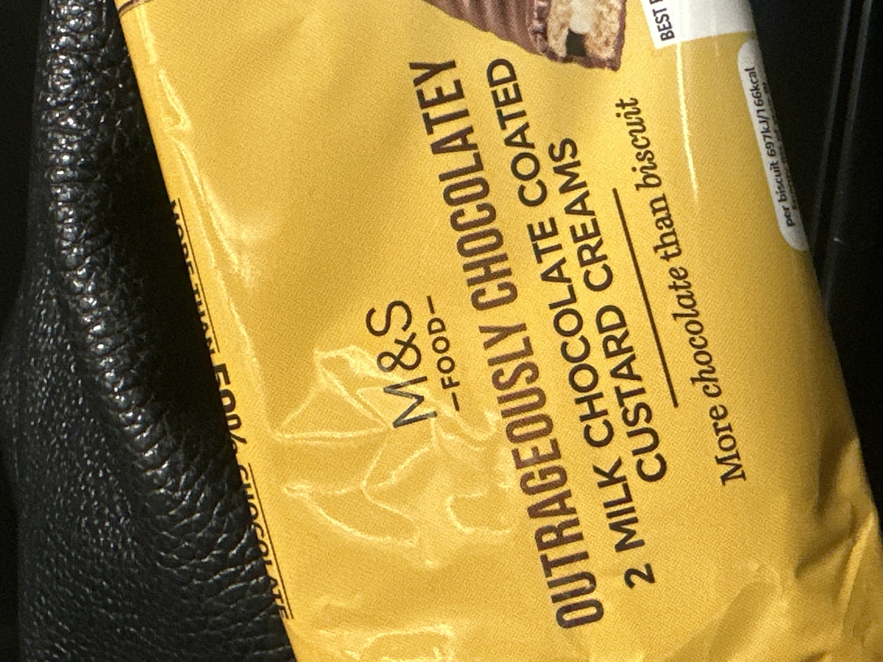 The image displays a close-up, angled view of a yellow food package, likely for biscuits or cookies, from the brand M&S. The package is positioned diagonally across the frame, with a dark, textured surface, possibly leather or faux leather, visible in the background on the left and right sides.
The dominant color of the packaging is a vibrant yellow, with a subtle grainy texture. Prominently featured on the package is the text: "M&S" in a large, stylized font, followed by "-FOOD-". Below this, in bold capital letters, is "OUTRAGEOUSLY CHOCOLATEY". Further down, it reads "2 MILK CHOCOLATE COATED CUSTARD CREAMS", with "MILK CHOCOLATE COATED" being larger than "CUSTARD CREAMS". Beneath this, a line separates the text "More chocolate than biscuit" in a cursive-like font.
Towards the top right of the package, a partial image of a biscuit is visible, showing a thick chocolate coating over a lighter, creamy or biscuit interior. Also visible on the right edge of the package, near the top, is the word "BEST" from what appears to be a "BEST BEFORE" date or label. Further down on the right side, nutritional information states "per biscuit 697kJ/166kcal".
The lighting suggests an indoor setting, with some glare or reflection on the plastic wrapping of the package, indicating it might be glossy. The dark background material has a distinct, small-scale ridged or bumpy pattern.