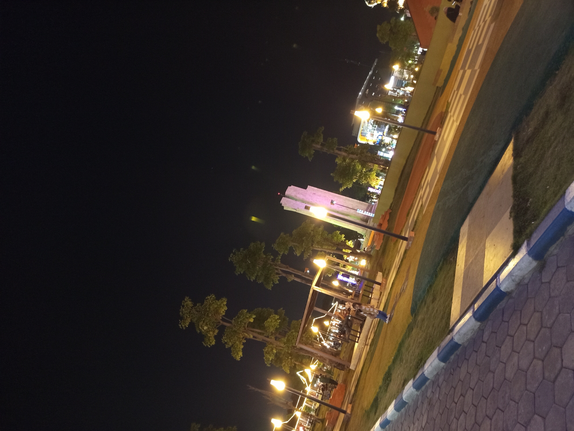 The image captures a nighttime scene at Alun-Alun Sidoarjo, a public square in Sidoarjo, Indonesia. The foreground features a paved walkway with hexagonal tiles and a blue and white striped border, leading into a grassy area illuminated by lampposts. In the mid-ground, several tall trees with sparse foliage are visible, along with an open-air pavilion or seating area. People are present, though mostly indistinct due to the low light conditions, with some appearing to be seated or walking. A prominent white, modern-looking monument or building with a pointed top stands to the left of the image's center, emitting a pinkish-purple light. Further in the background, several buildings with illuminated windows suggest commercial or public structures. The overall atmosphere is that of a lively urban park at night, with artificial lighting creating a warm glow.