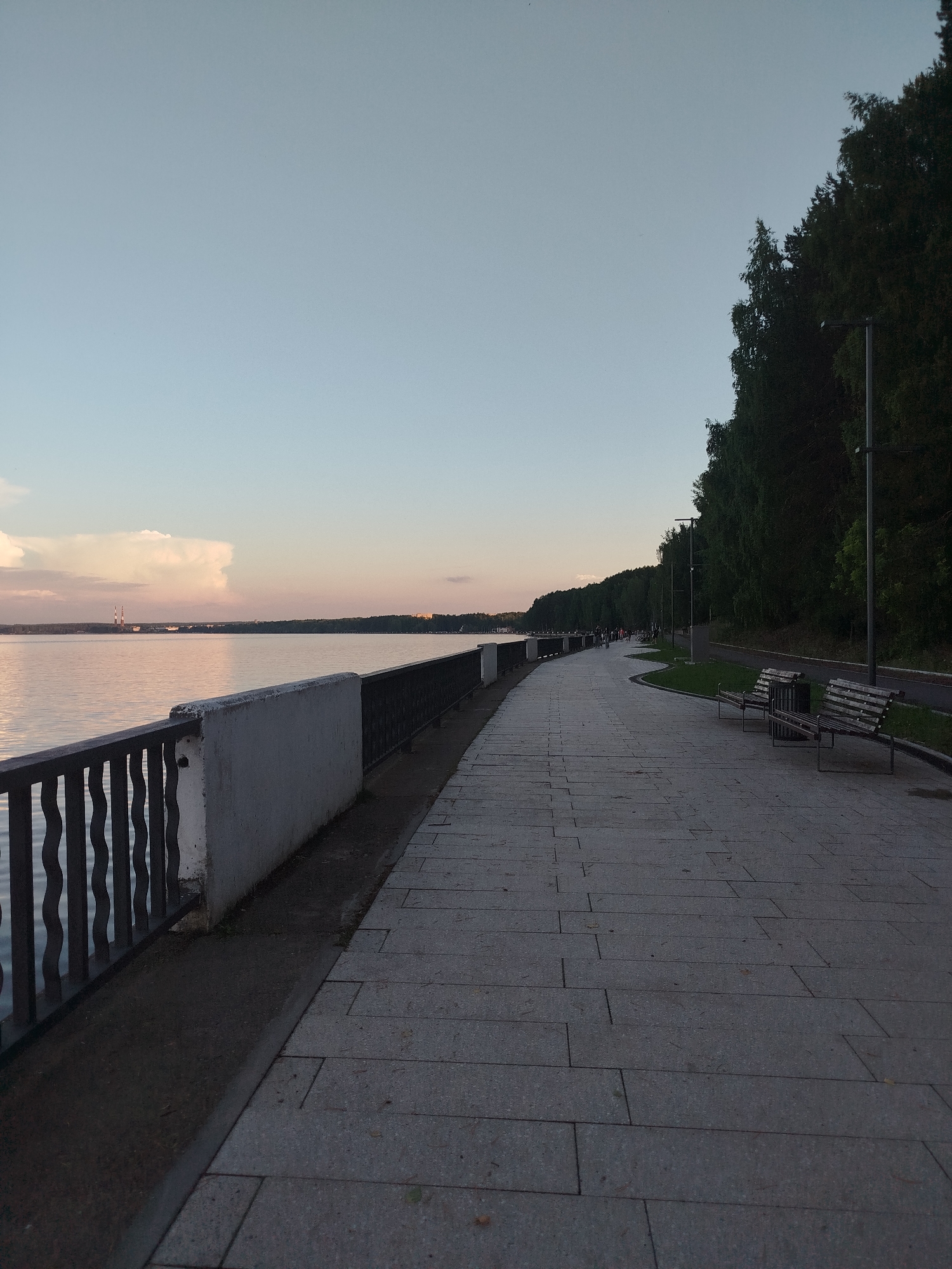 An outdoor, eye-level, medium shot captures a paved embankment along a large body of water, likely a river or lake, under a pale, dusky sky. The sky transitions from a pale blue at the top to a soft pink and orange near the horizon. The water is calm and reflects the muted colors of the sky, with a few distant, silhouetted industrial structures and clouds visible on the far shore.
The embankment itself is wide and paved with large, rectangular grey stones. A dark metal railing with a decorative, wavy pattern runs along the edge of the water, separated from the path by a white concrete barrier topped with more dark metal railing. The embankment curves gently to the right, leading the eye into the distance.
To the right of the paved path, a strip of green grass is bordered by a paved walkway, likely for cycling or jogging. Several dark metal lampposts with a modern design are spaced along this walkway. Two wooden park benches with metal legs are positioned on the grass, facing the water.
Further down the embankment, a line of dense, dark green trees forms a backdrop, their leaves appearing dark against the twilight sky. Scattered figures of people are visible walking and standing along the embankment, appearing as small silhouettes. The overall impression is one of a serene, public recreational area at dusk.