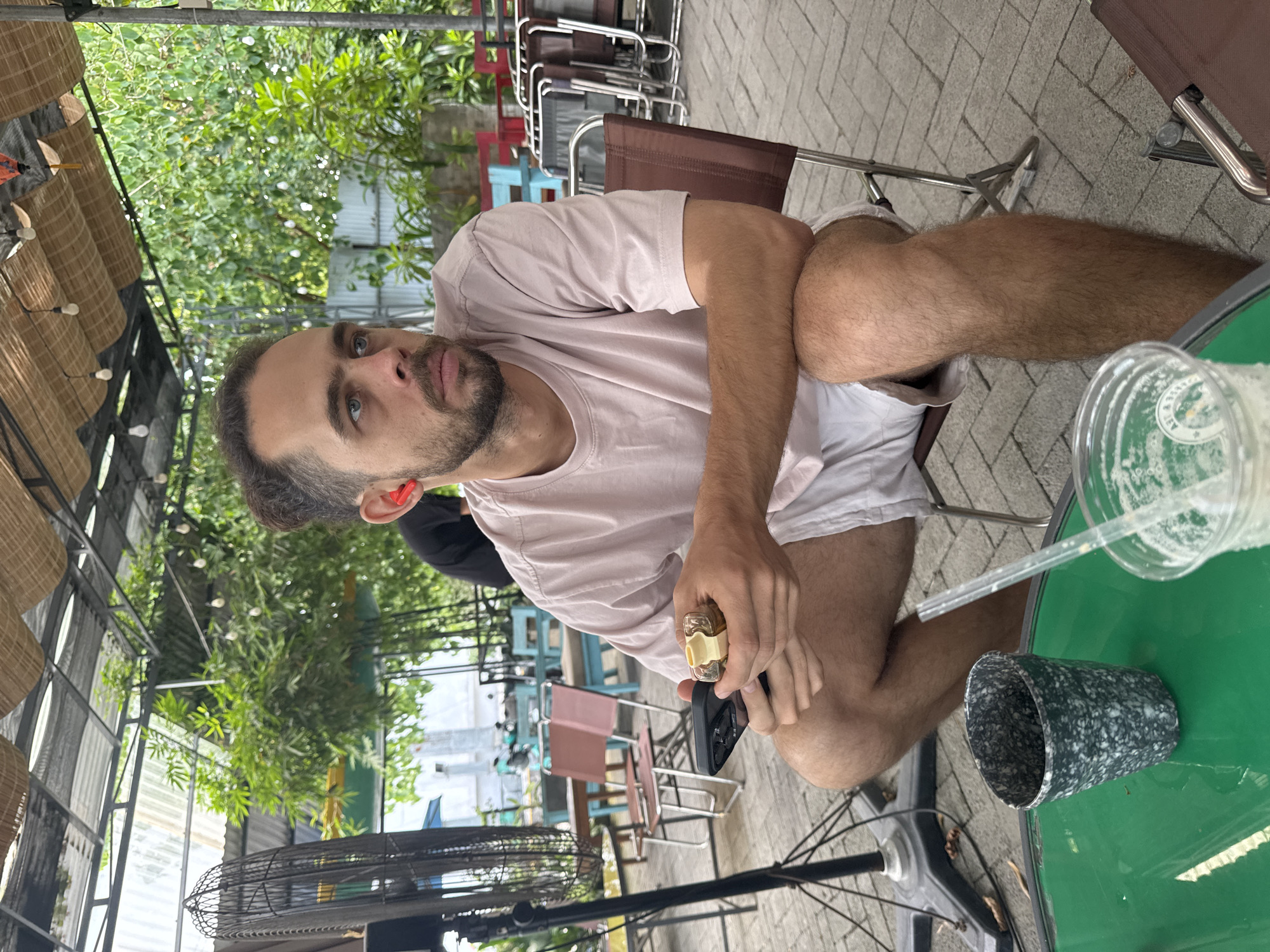 A man sits outdoors at a cafe, holding a phone and spray. The location is potentially Đà Nẵng, Vietnam, suggested by the building style and tropical vegetation in the background. The man, with a beard, is wearing a light pink t-shirt and white shorts. He has a drink on the green table. A fan can be seen in the background.