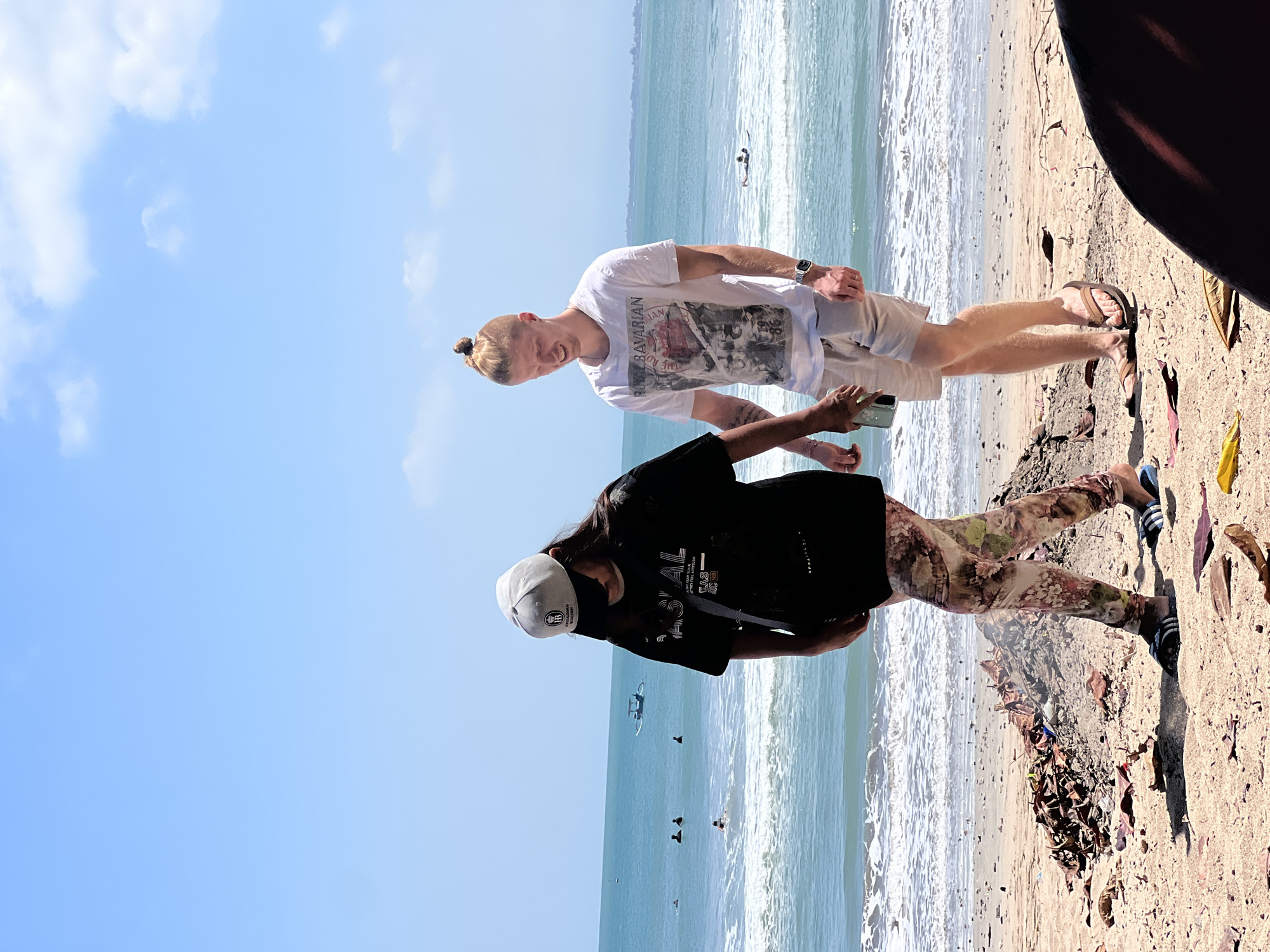 The image shows a beach scene in Kuta, Indonesia, on a bright, sunny day. Two people, a woman wearing a black t-shirt, a grey baseball cap, and floral-patterned leggings, and a man in a white t-shirt with graphics and beige shorts, are walking side-by-side on the sand. The woman is looking down, and the man is looking forward with a slight smile. They are walking towards the right side of the frame.
In the background, the turquoise ocean stretches out, with gentle waves breaking on the shore. Several people are visible in the water; some are swimming, and one person appears to be on a surfboard. A small boat is also visible further out at sea. The beach itself is sandy, with some debris, including dried leaves and small piles of what appears to be burnt material, scattered on the ground. The sky is a clear blue with a few white clouds. The lighting suggests it is daytime.