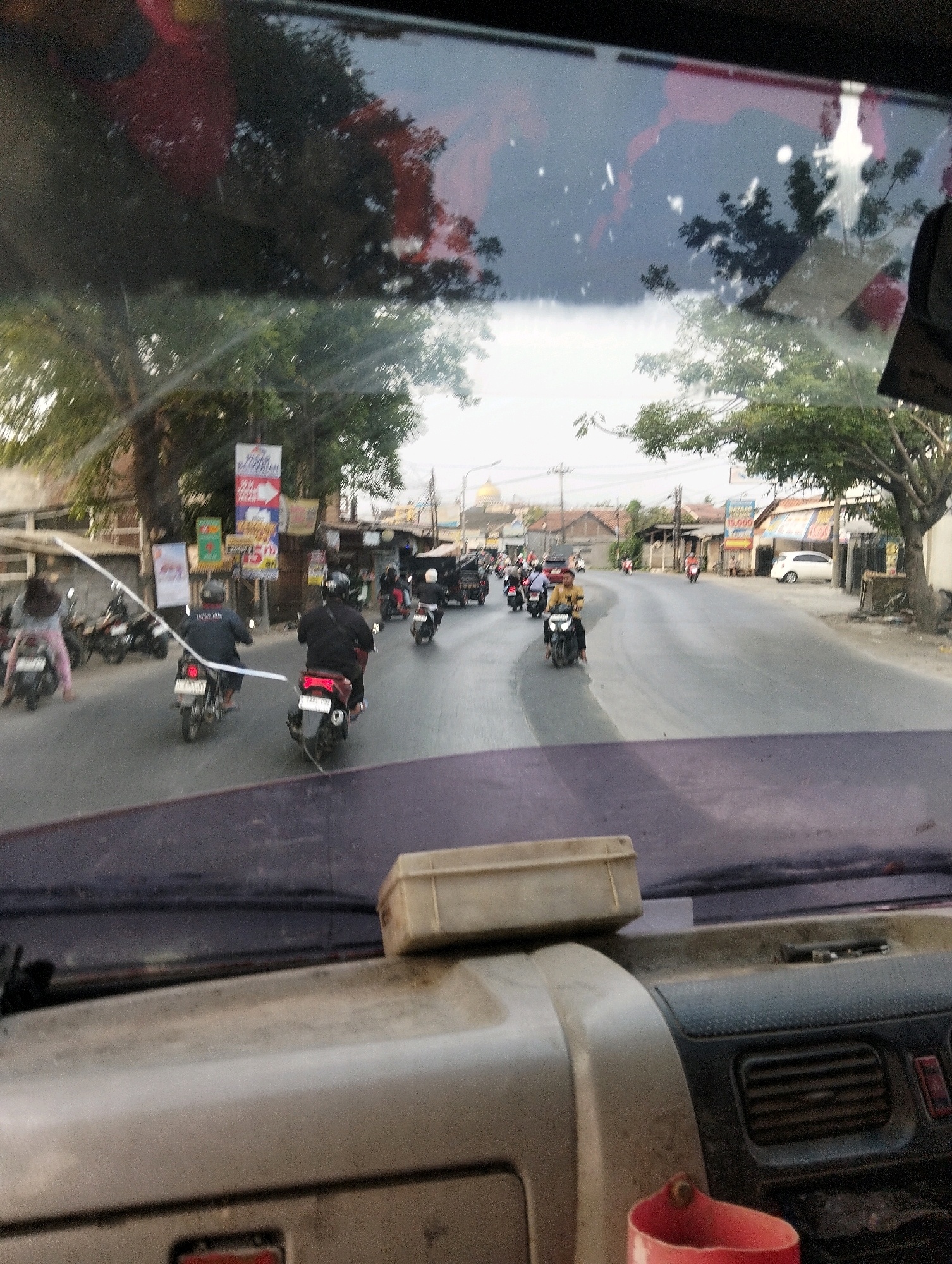 The image depicts a street scene in Semarang, Indonesia, viewed from the interior of a vehicle through the windshield. The road is paved and appears to be a typical urban street with buildings lining one side. Numerous motorcycles and a few cars are present on the road, indicating moderate traffic. Several people are riding motorcycles, some wearing helmets. The environment suggests a warm climate with trees visible along the roadside and on the horizon. The sky is overcast, with hints of sunlight breaking through. The foreground shows the dashboard of the vehicle, including a container and what appears to be part of a cup holder. The overall impression is of a busy street in an Indonesian city.