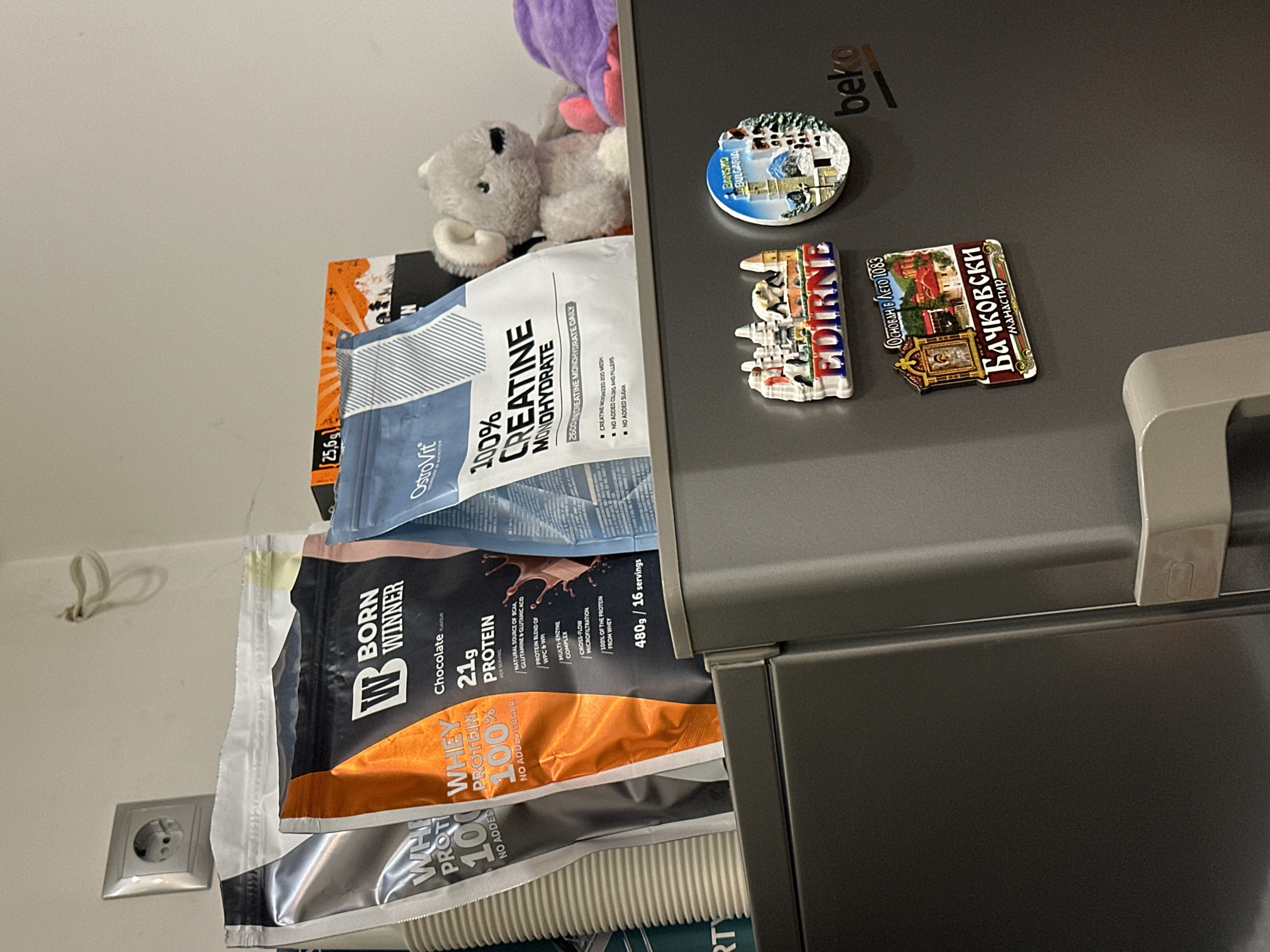 The image shows a cluttered scene near a refrigerator. On top of the refrigerator, there is a plush purple toy and a grey teddy bear. Several items are stacked or leaning against the side of the fridge. These include a blue and white package of "OstroVit 100% CREATINE MONOHYDRATE" and a black and orange package of "BORN WINNER WHEY PROTEIN 100%," with nutritional information visible.
The refrigerator door has several magnets on it. These include a magnet depicting Bansko, Bulgaria, another of Edirne, and one of the Bachkovo Monastery (Бачковски манастир), which was established in 1083.
Visible in the background is a white wall with an electrical outlet.