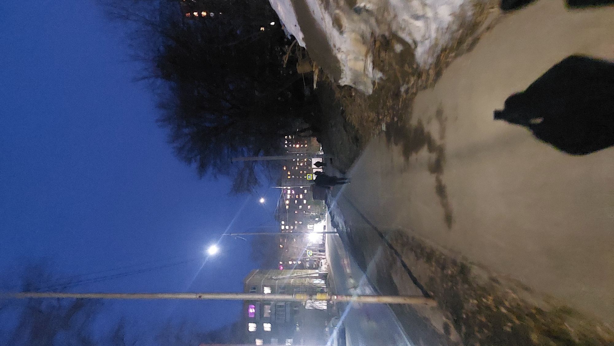 The image presents a nighttime scene from a street in Novosibirsk, Russia. A sidewalk runs along the right side of the image, slightly elevated from the street, with some snow and debris along the edge. A solitary figure and another individual or couple walk along the sidewalk in the middle distance. Streetlights illuminate the area, casting shadows and reflections on the wet pavement. In the background, multi-story apartment buildings with illuminated windows stretch into the distance. A bare tree stands on the left side of the street, reaching up into the dark blue night sky. The perspective is distorted, possibly due to the camera angle.