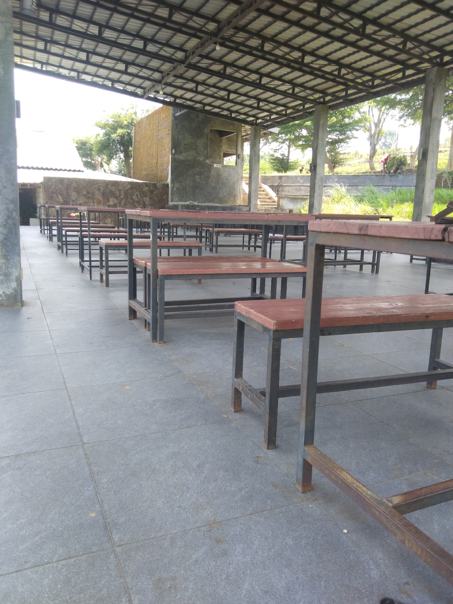 The image depicts an outdoor covered seating area with multiple rows of wooden benches and tables with metal frames. The flooring is made of grey square tiles. In the background, there are trees, a stone wall, and a concrete structure that appears to be under construction or partially built, with visible rebar. Sunlight is filtering through the roof structure. Further in the background, two individuals are visible near a grassy embankment, one carrying a load of grass. The location is identified as Keling, Indonesia.