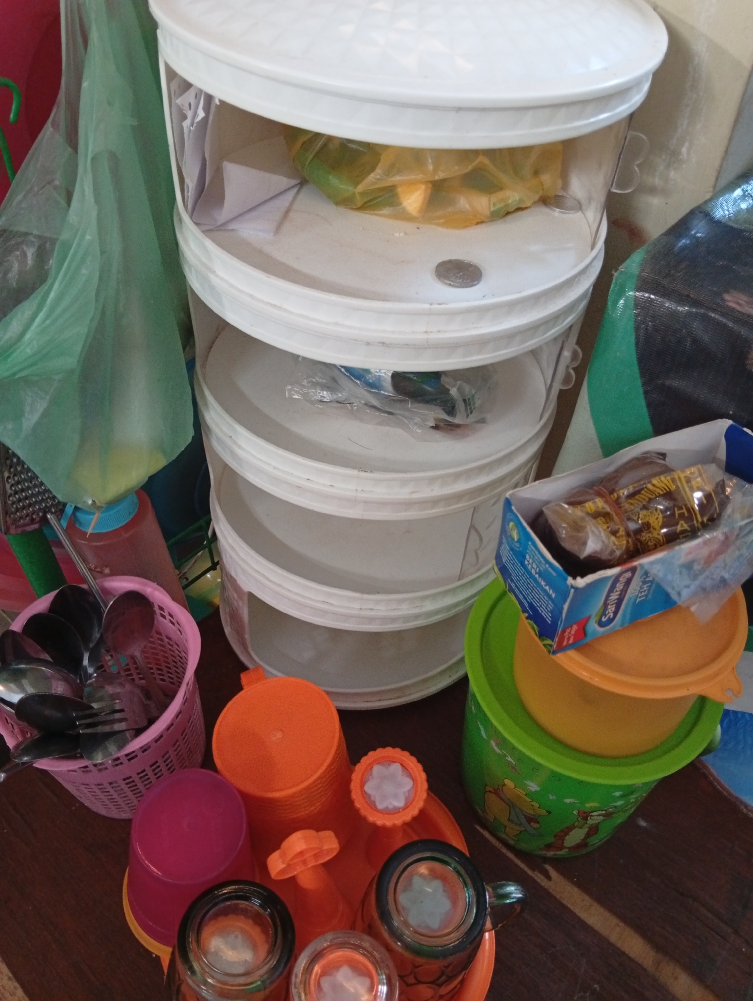 The image depicts a cluttered indoor scene, likely a kitchen or storage area, with various household items. In the center is a four-tiered white plastic organizer with clear sections, each containing some contents, possibly food items in plastic bags on the upper shelves. To the left, a green plastic bag hangs, and a red and blue squeeze bottle is visible. A pink basket filled with spoons and forks sits on a wooden surface. In the foreground and right side, several plastic containers and cups are stacked and arranged. A green bucket with images of Winnie the Pooh characters is prominent. A blue and white cardboard box, likely containing tea packets, rests on top of an orange lid. The overall impression is one of disorganization and everyday clutter. This scene is located in Kualu, Indonesia.