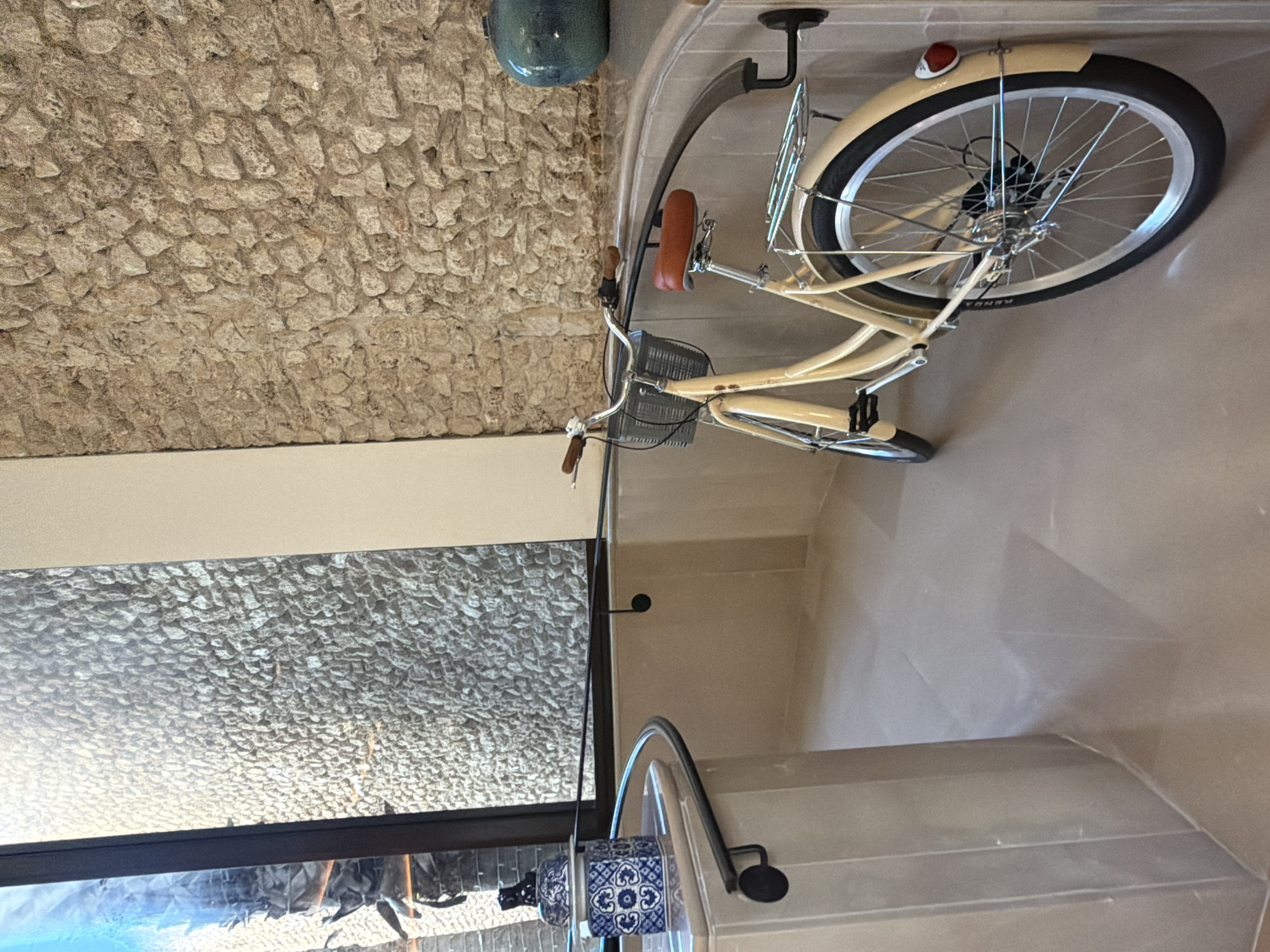 The image shows a bicycle hanging on a wall indoors. The bicycle is light tan with a brown seat and a front basket. The wall behind the bicycle is made of rough, light-colored stone. A glass partition is visible, with a view to the outside. A decorative object with blue and white patterns sits near the bottom of the glass partition. The location appears to be indoors, possibly a residence in Tel-Aviv, Israel.