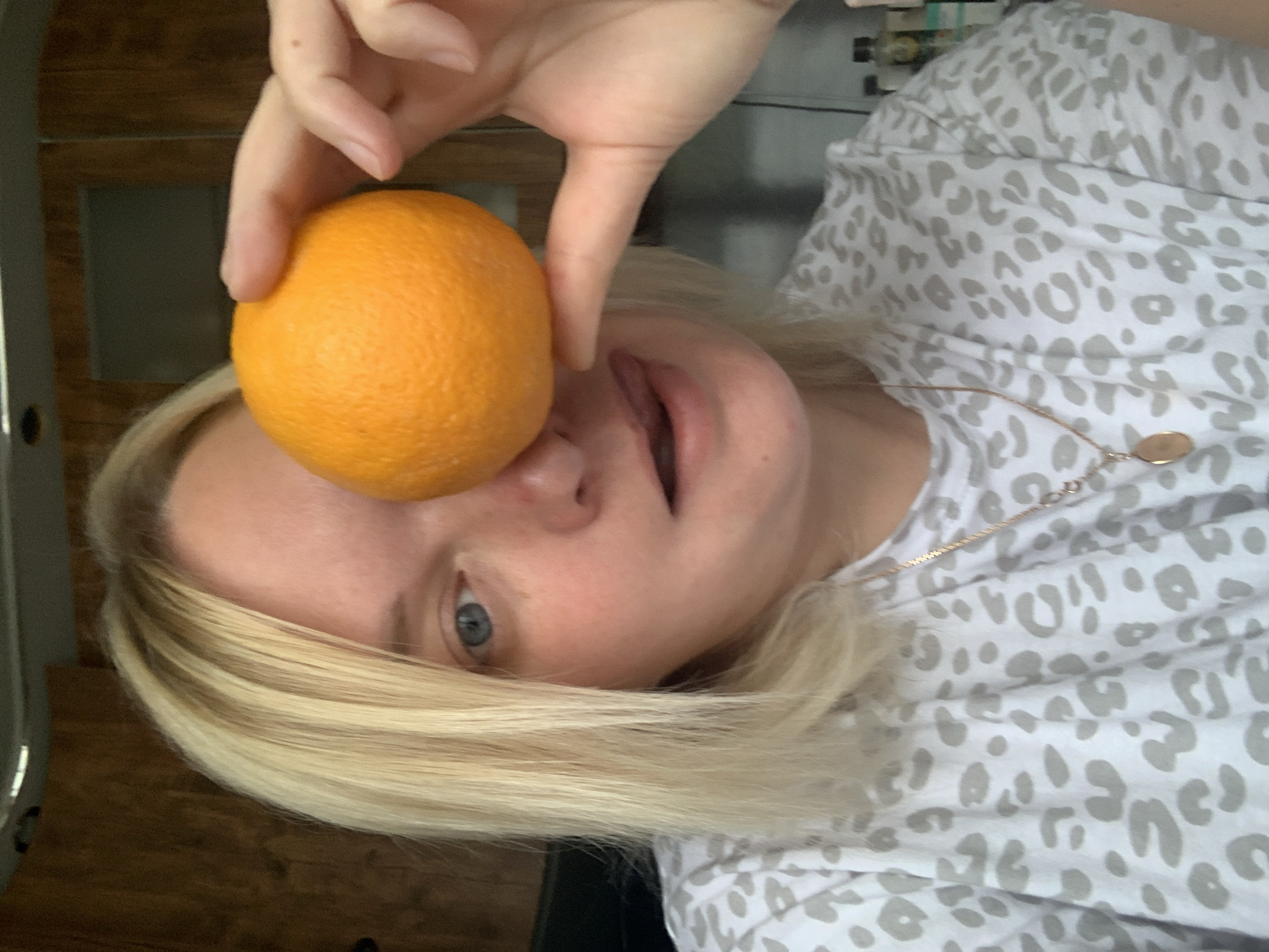 An indoor, close-up, and slightly rotated portrait captures a person holding an orange on their face. The subject is a fair-skinned individual with light blonde hair, which frames the left side of their face. One blue eye is visible, looking towards the viewer, while the mouth is slightly open, suggesting a playful or surprised expression. A single, whole, ripe orange is held by the person's fingers, resting directly on their forehead and bridge of the nose, partially obscuring the upper part of their face.
The person is wearing a white t-shirt adorned with a light grey leopard print pattern. A delicate, gold-colored chain necklace with a small, round, flat pendant is visible around their neck, resting on the shirt. The background suggests a home interior, with dark brown wooden panels or furniture visible on the left and in the lower right corner, indicating a warm, natural wood tone. Other elements in the upper background are indistinct and dark, possibly shelves or appliances. There is no visible text in the image. The overall mood appears lighthearted and spontaneous. The given location context is Chaikovsky, Russia.