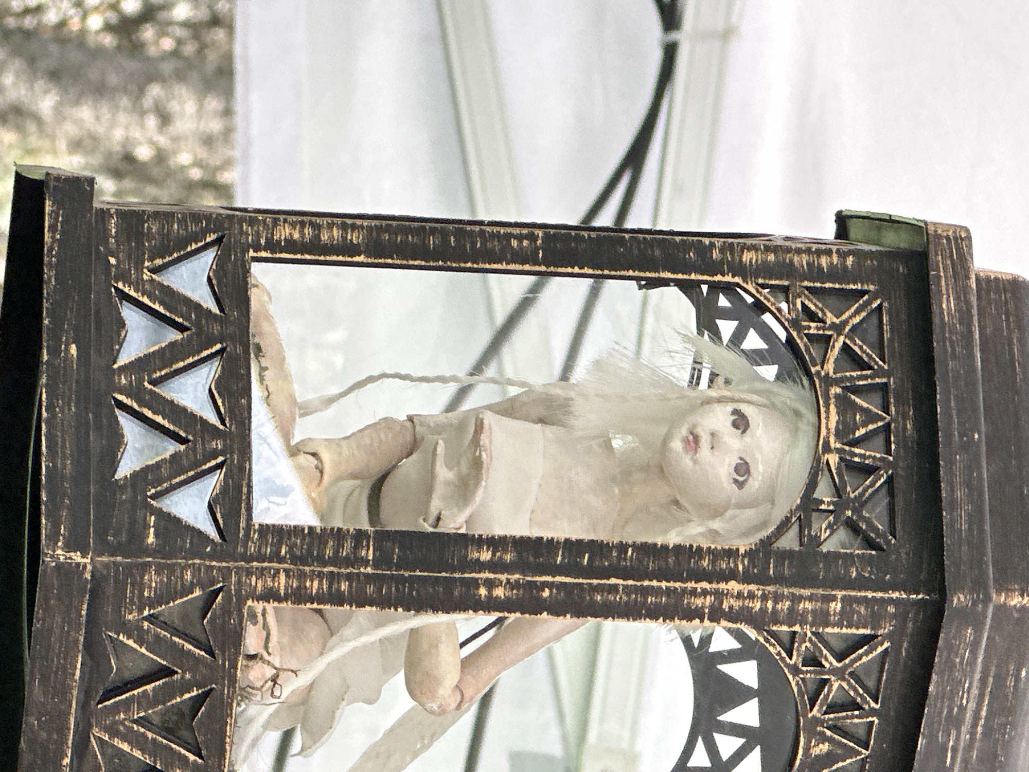A close-up shot depicts a porcelain doll encased within a decorative metal frame, likely part of an outdoor art installation. The frame is dark, possibly bronzed, with geometric cut-out patterns and what appears to be glass or reflective material within the openings. The doll has pale skin, light hair, and dark eyes; it is partially wrapped in a light-colored fabric, giving it an ethereal or otherworldly appearance. The environment around the frame suggests an outdoor setting, with blurred foliage and glimpses of what might be a white tent in the background. The location is not discernible.