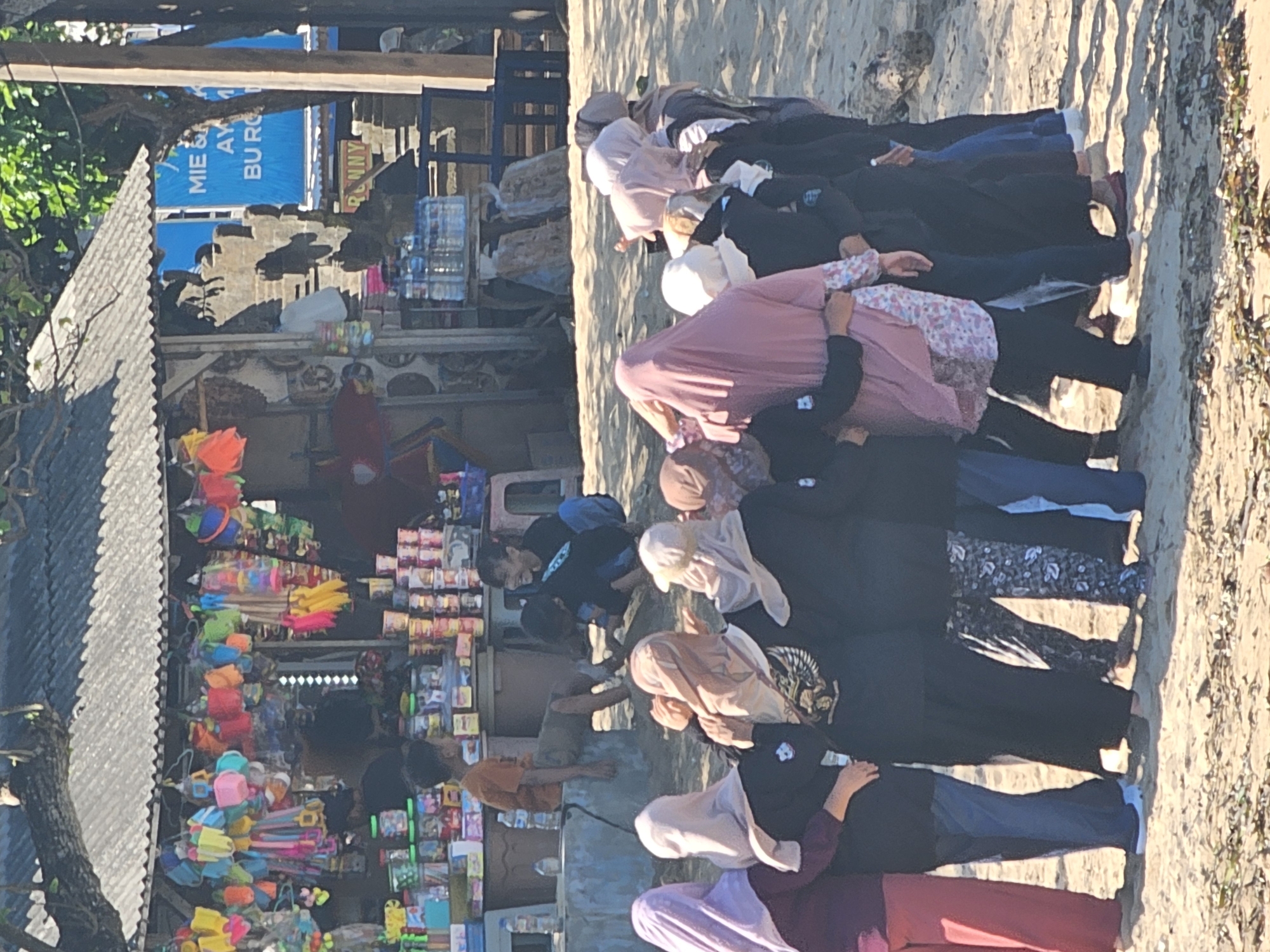 In Denpasar, Indonesia, on a sandy beach, a group of individuals, predominantly young women wearing headscarves, are gathered in a circle, their arms interlocked. They face inwards, suggesting a moment of shared activity or connection. In the background, a small open-air stall displays an array of colorful merchandise, including bags, toys, and snacks, indicating a tourist or local market area. Two young boys are visible near the stall, one sitting and the other kneeling, interacting with the merchandise or their surroundings. The scene is set under the bright sun, with dappled shadows suggesting trees or structures nearby. The overall atmosphere appears casual and relaxed, set against the backdrop of a beach and a commercial stall.