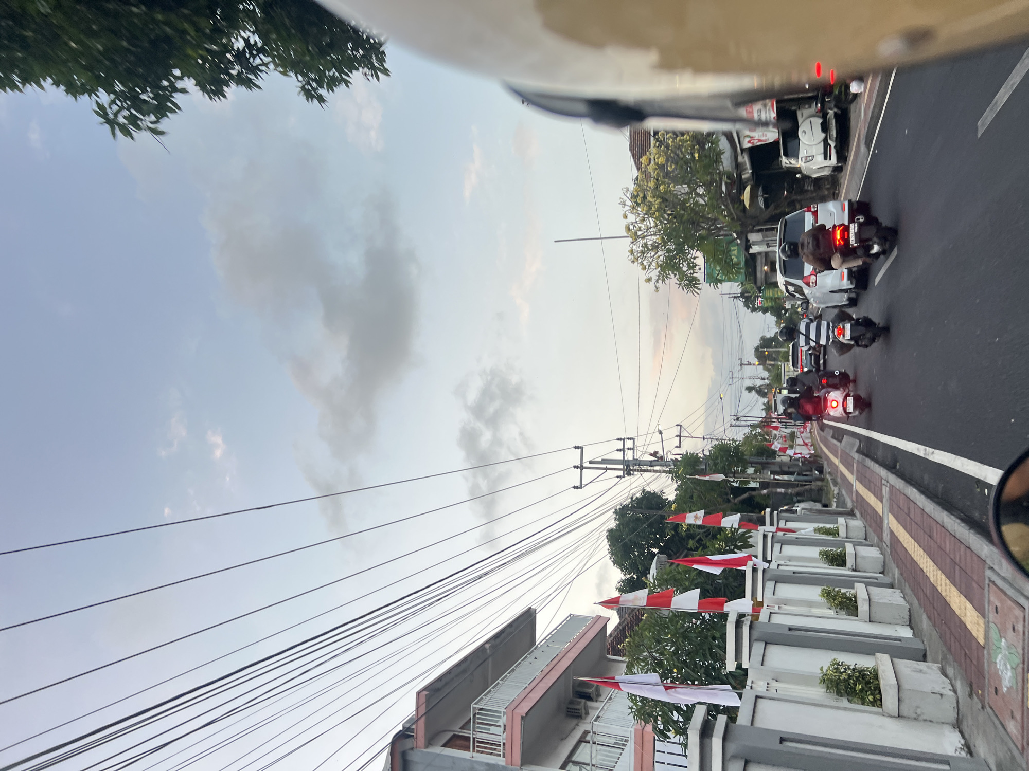 The image captures a street scene in Denpasar, Indonesia, likely during a festive occasion given the presence of numerous Indonesian flags displayed along a sidewalk. The perspective is from the viewpoint of someone on a motorcycle, looking forward. The road is moderately busy with traffic, featuring several motorcycles and cars. On the left side of the road, a building with multiple balconies is visible, adjacent to a neatly maintained sidewalk with planters and red and yellow patterned paving stones. The sidewalk is lined with flags. The right side of the road shows a row of shops and businesses. Overhead, a network of power lines crisscrosses the sky. The sky itself is a mix of blue and grey clouds, suggesting it might be late afternoon or early evening. The overall atmosphere appears to be that of a typical urban street in Denpasar.