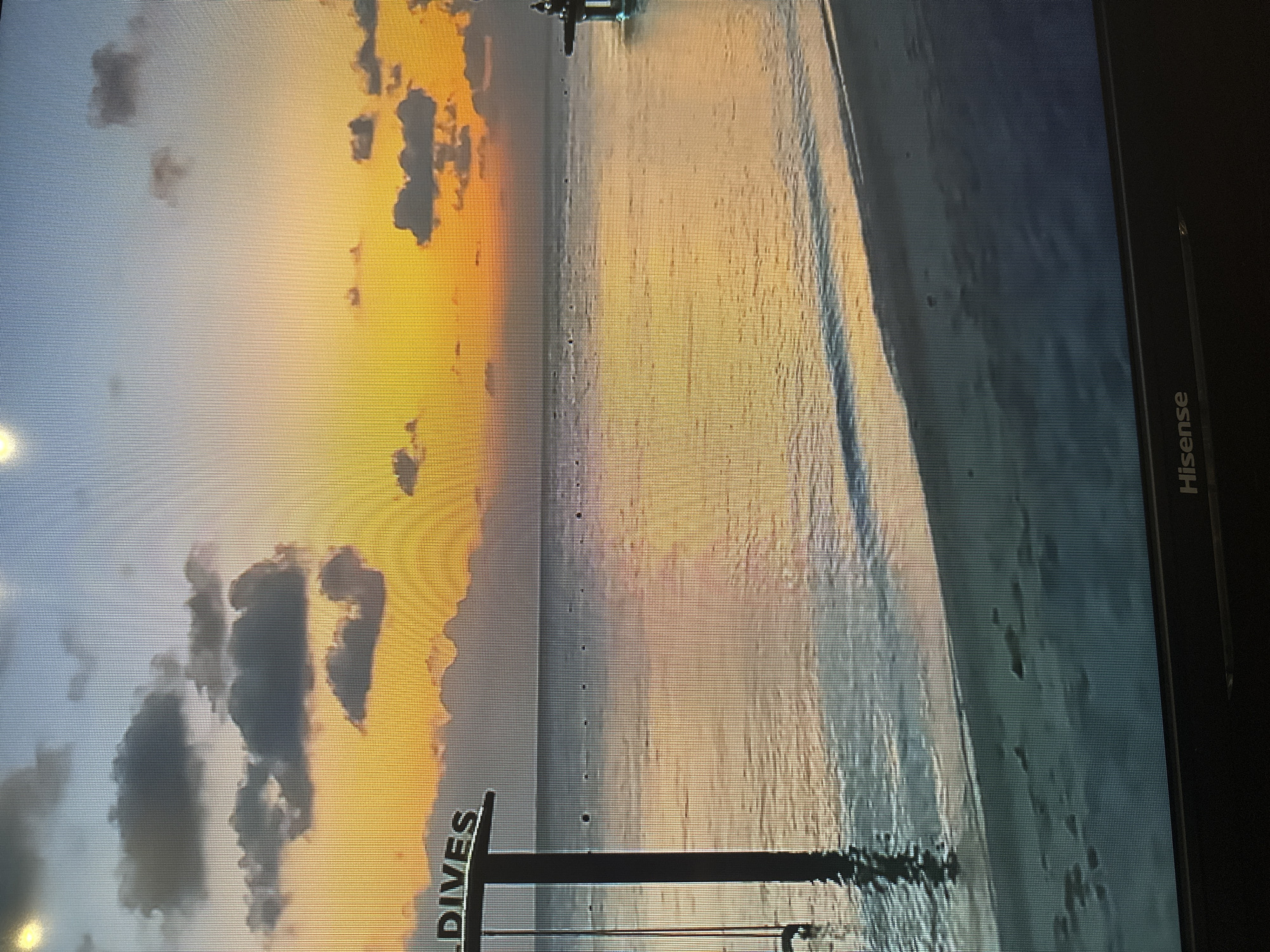 The image depicts a tranquil beach scene at sunset. The sky is a gradient of warm colors, transitioning from a bright yellow and orange near the horizon to a soft blue at the top, with scattered dark clouds. The calm ocean reflects the colors of the sky, creating a shimmering expanse of water. A sandy beach is visible in the foreground, with footprints imprinted on the wet sand.
On the left side of the image, a dark pole stands partially submerged in the water, with the word "MALDIVES" visible at the top. This suggests a tropical resort or island destination. In the distance, small buoys dot the water, and a pier-like structure can be seen on the right.
The bottom of the image shows a black Hisense television screen. The image itself is displayed on this screen. There is no discernible indication that this scene is located in Kyiv, Ukraine. The visual elements strongly suggest a tropical beach environment.