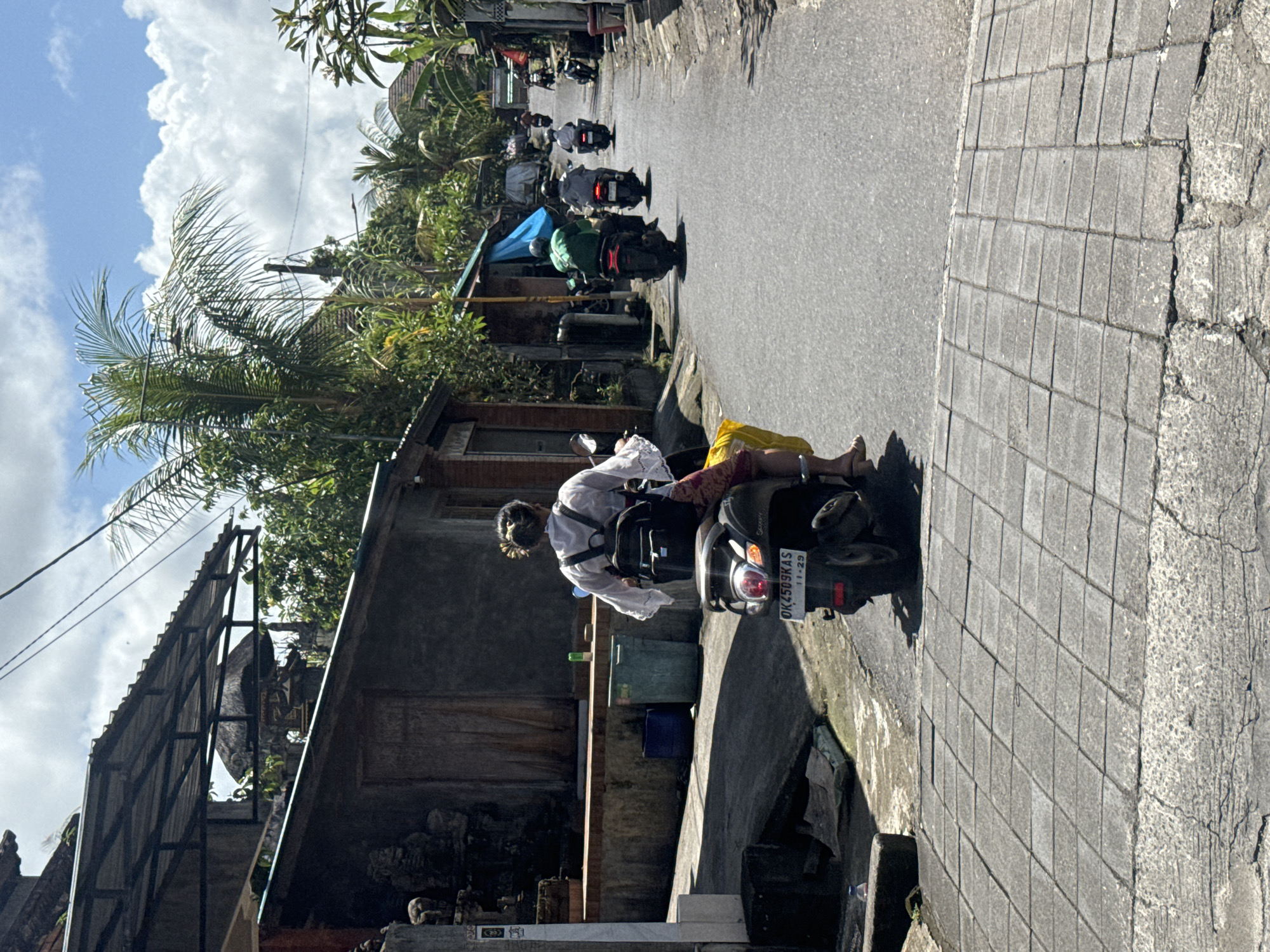 The image shows a street scene in the Ubud District of Indonesia. A woman in a white shirt and yellow pants is riding a black scooter with the license plate "DK 4509 KAS". The scooter is traveling on a narrow asphalt road with a concrete sidewalk on one side. Other scooters are visible in the distance, moving in the same direction. The scene is lined with buildings, trees, and greenery typical of the Ubud area. The sky above is bright with scattered clouds.