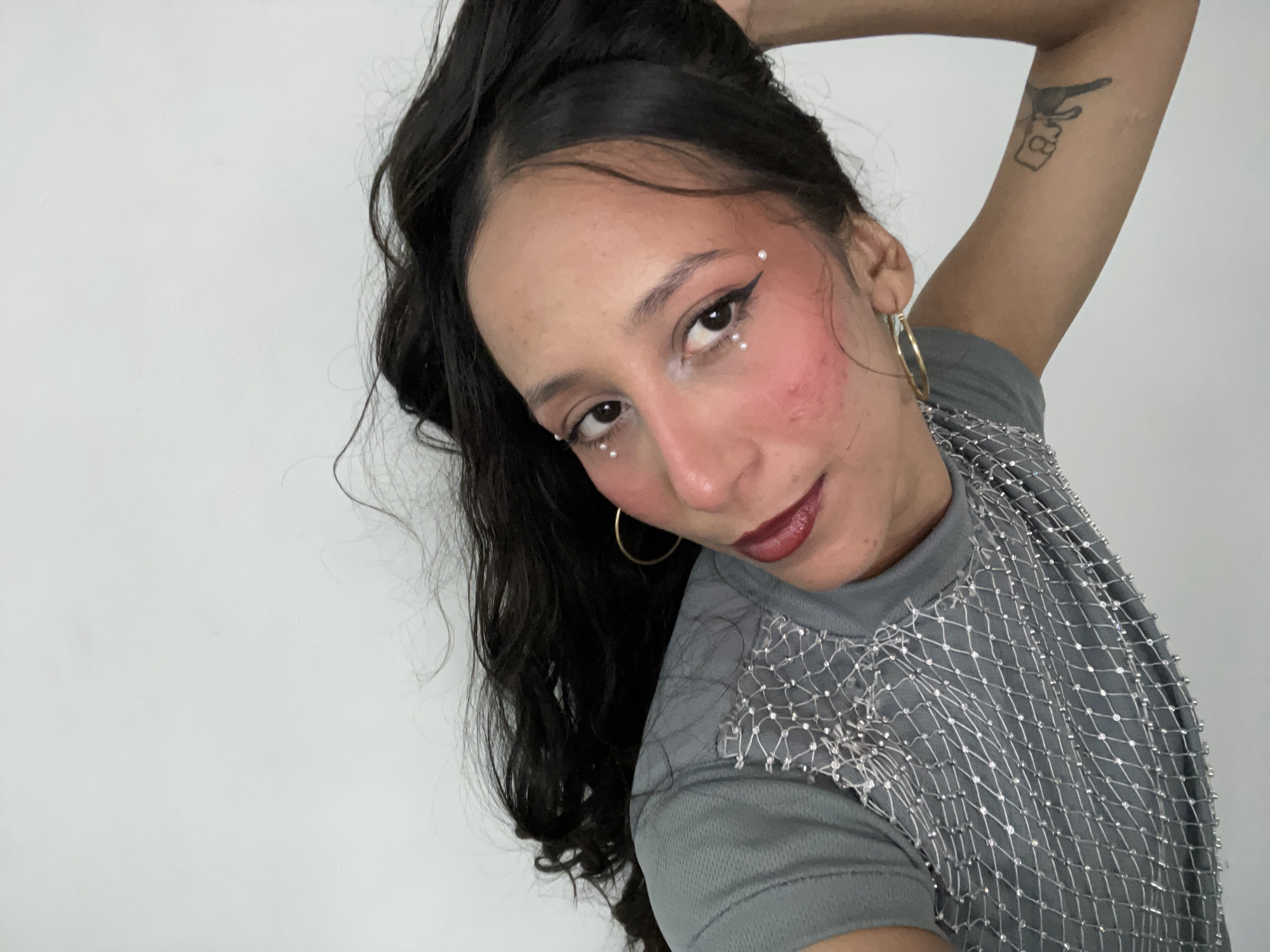 A young woman with dark hair and olive skin is positioned in the foreground of the image, looking towards the camera with a slight smile. Her hair is pulled up, with some strands falling loosely around her face and shoulder. She is wearing makeup, including winged eyeliner, blush, and lipstick, with small white pearls adorning her eyelids and under her eyes. She has gold hoop earrings and a tattoo on her upper arm, depicting what appears to be a hand holding a gun with the number "8" within a box. Her attire consists of a grey t-shirt with a translucent, net-like overlay embellished with small silver beads across the shoulders and upper chest. The background is a plain white wall. The scene does not contain any discernible elements indicating its location within Porlamar, Venezuela.