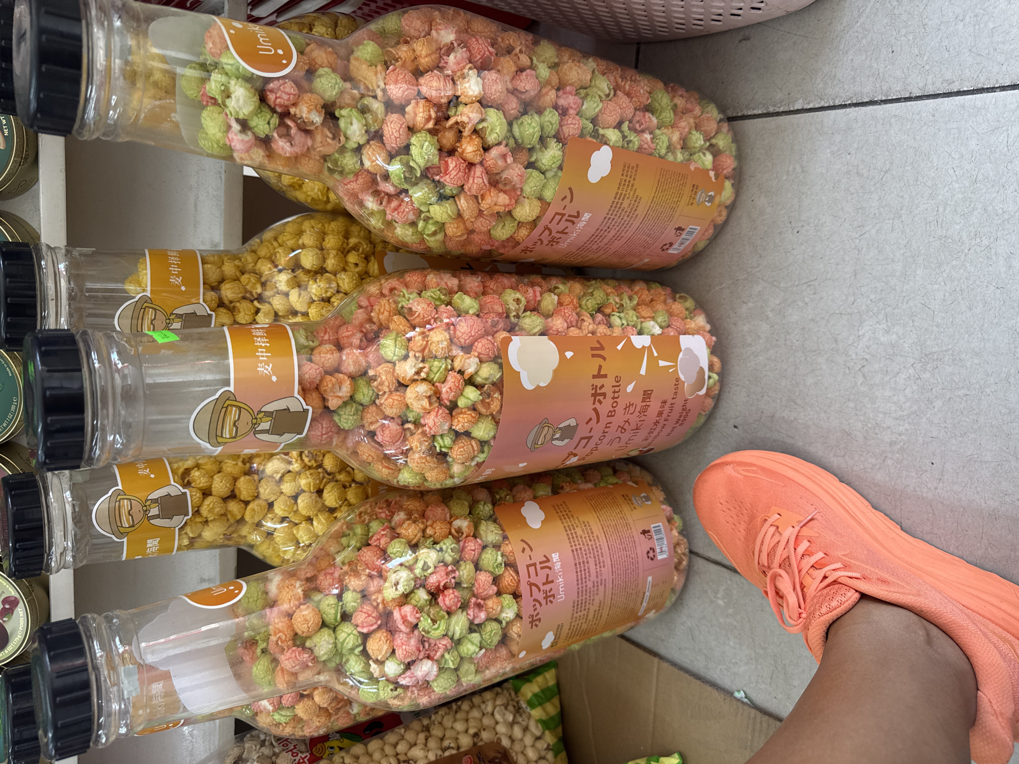 The image depicts a close-up, ground-level view of several large, clear plastic bottles filled with colorful popcorn. The bottles are arranged in a diagonal line, with the one closest to the viewer on the left. The popcorn inside is a mix of orange, pink, and green kernels. Each bottle has a label with Japanese text and illustrations of a cartoon character wearing a hat and apron. The bottle on the far left is partially obscured by the foot and lower leg of a person wearing a bright orange athletic shoe. The background suggests a retail environment, with more bottles and shelves visible. The floor appears to be tiled. The context of Nha Trang, Vietnam, is not discernible from the visual information provided in the image.