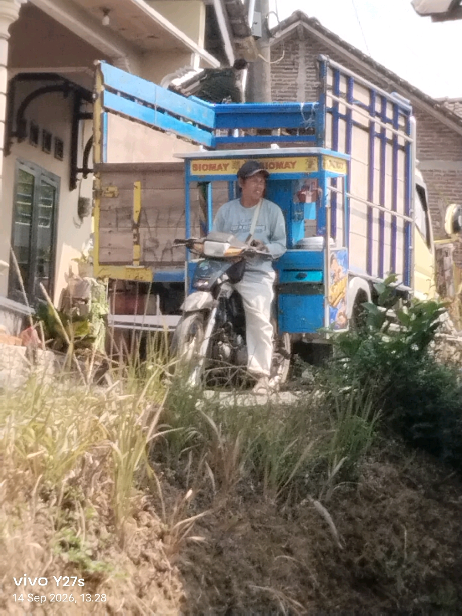 A man wearing a light blue shirt, dark baseball cap, and white pants stands next to a motorcycle. He is holding a paper and appears to be looking to his left. The motorcycle is parked next to a blue and yellow truck with the word "SIOMAY" visible on the side. The truck is parked on a dirt road, with tall grass and vegetation in the foreground. In the background, a building with a tiled roof can be seen. The scene appears to be in a rural or suburban area. The timestamp indicates the image was taken in Tamansari, Indonesia on September 14, 2026, at 13:28.