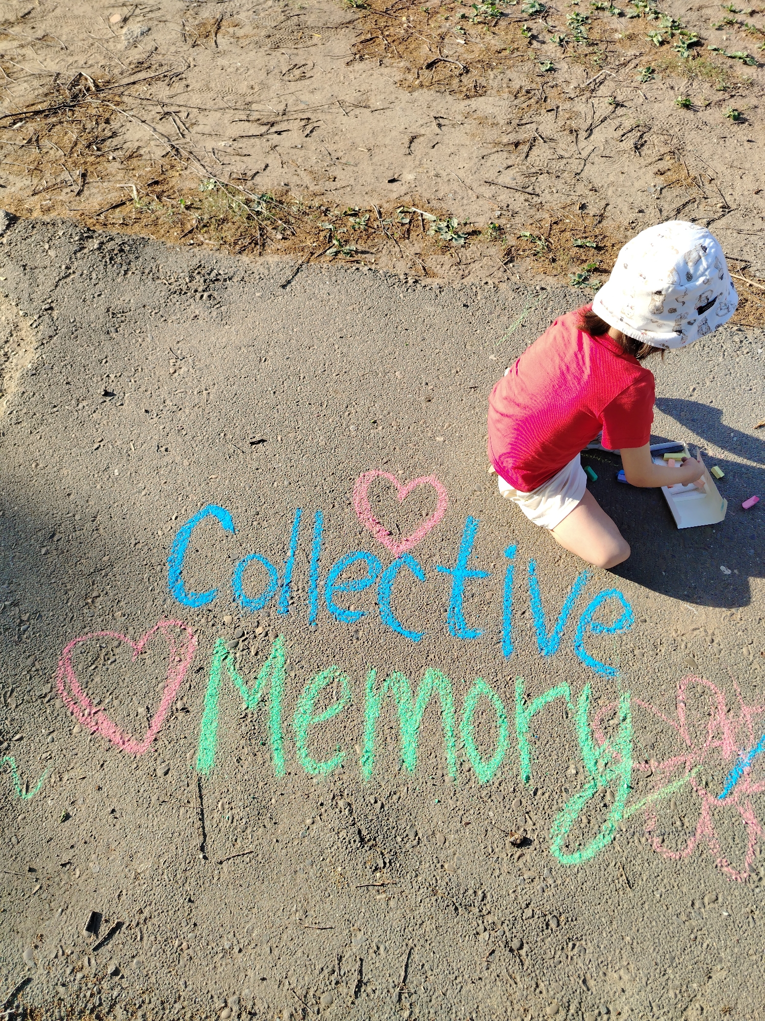 A child in a red shirt and white shorts kneels on a paved surface drawing with chalk. The pavement is light gray and abuts an area of dirt and sparse vegetation. The words "Collective Memory" are written in chalk, with "Collective" in blue, "Memory" in green, and heart shapes in pink on either side. The child wears a white hat with a pattern. A white box of chalk sits next to the child, with various colored chalk sticks scattered around.