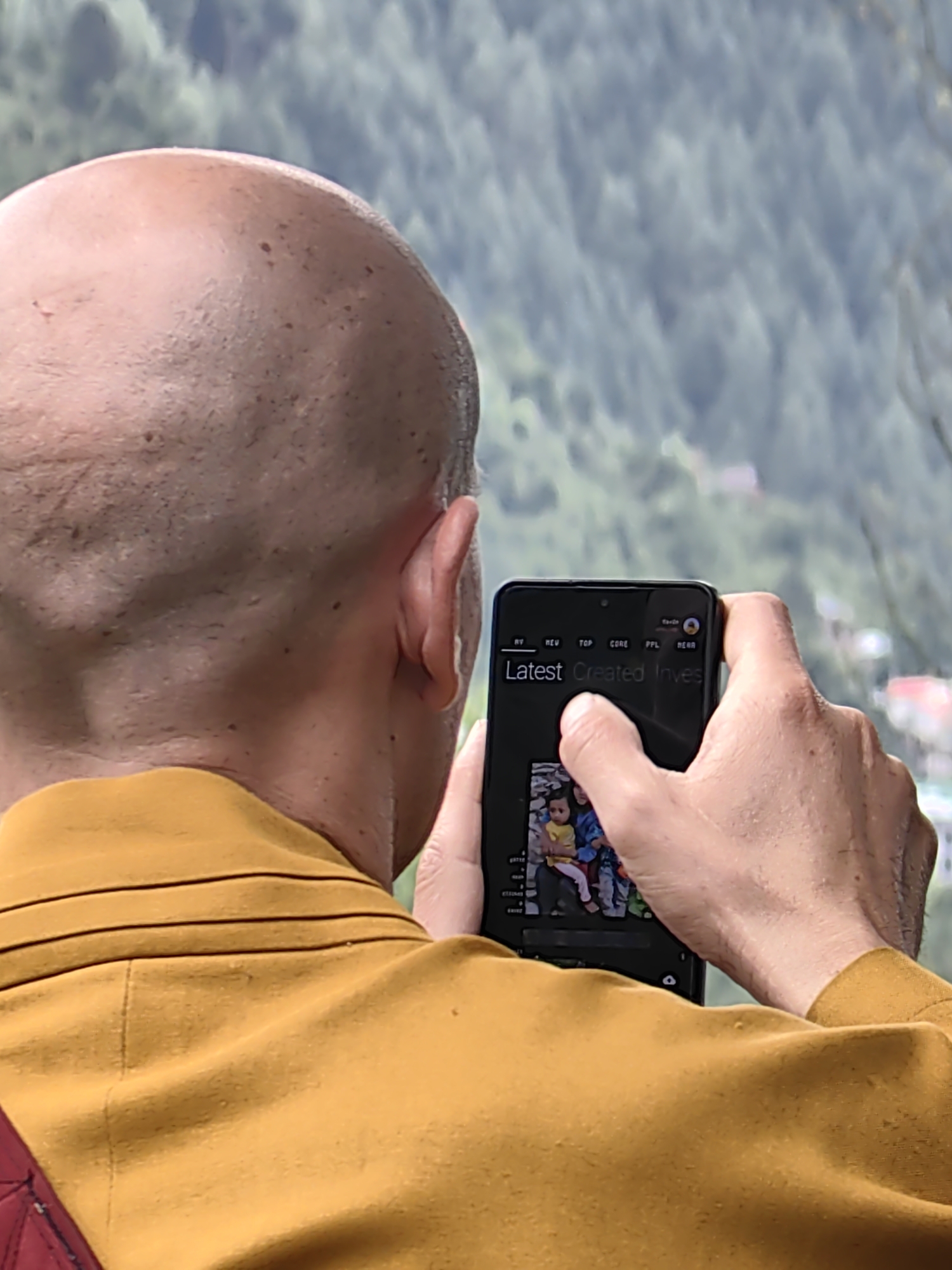In Vashisht, India, a bald individual wearing a mustard-yellow robe is captured from behind, holding a black smartphone to take a photograph. The person's right hand is visible, with fingers extended to operate the phone's touchscreen. The phone displays a vertical image featuring two children, one in a yellow shirt and the other in blue, seated together. Above the image on the phone, text reads "Latest Created Inves" with other labels such as "RY", "REV", "TOP", "CORE", "PPL", and "HERR". The background consists of a densely forested hillside, suggesting a natural, possibly mountainous, environment outside a city. The lighting appears to be natural daylight, casting subtle shadows on the person's skin and clothing.