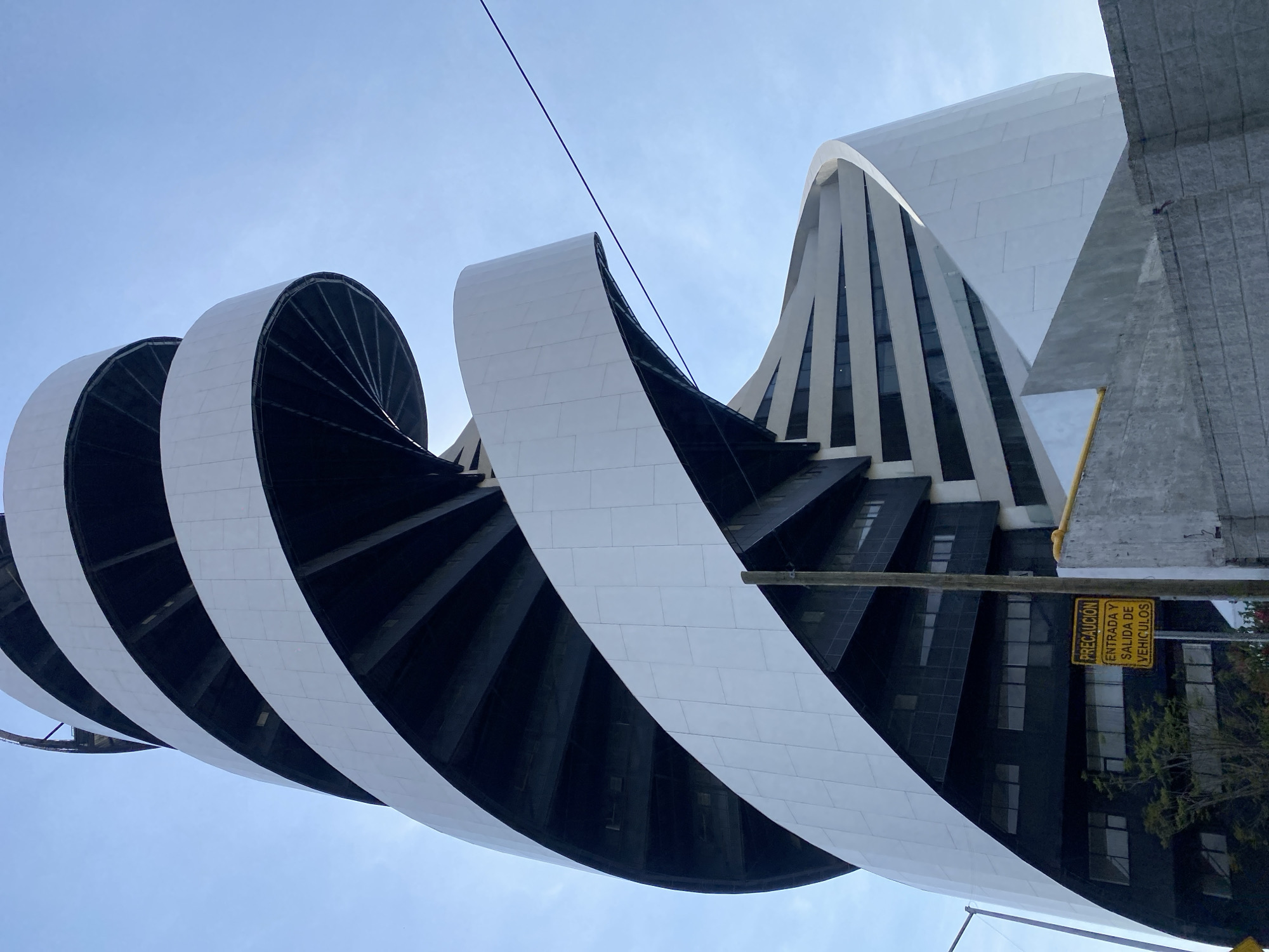 The image shows a unique architectural structure in Unknown, Unknown. The building's design is characterized by a series of curved, white-tiled elements that spiral around a darker, presumably black, inner structure. This creates a visually striking, almost sculptural effect.
Adjacent to this distinct structure is another building, featuring a pattern of alternating light and dark vertical stripes, likely indicating windows and supporting architectural elements. A yellow sign, with Spanish text "PRECAUCIÓN ENTRADA Y SALIDA DE VEHICULOS," is visible. The sky is clear and light blue, providing a backdrop to the buildings. A cable or wire stretches across the upper portion of the image.