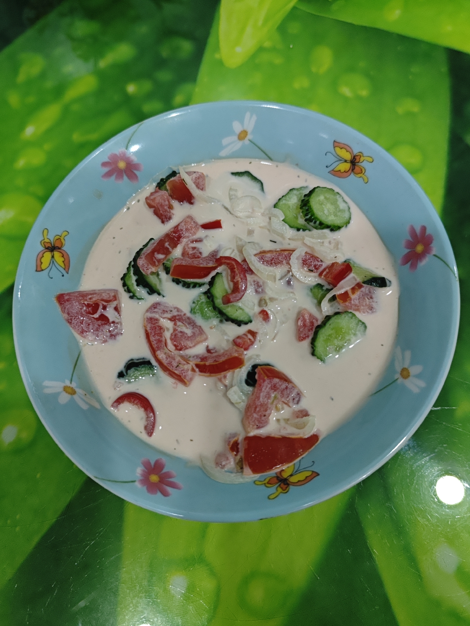 A light blue ceramic plate with a floral and butterfly pattern is filled with a creamy salad. The salad consists of chopped red tomatoes, sliced green cucumbers, and thinly sliced white onions, all coated in a white dressing. The plate rests on a green surface with a textured, almost leafy pattern, possibly a tablecloth or placemat. The lighting suggests an indoor setting, with a bright reflection on the green surface. No people or specific location details are discernible.