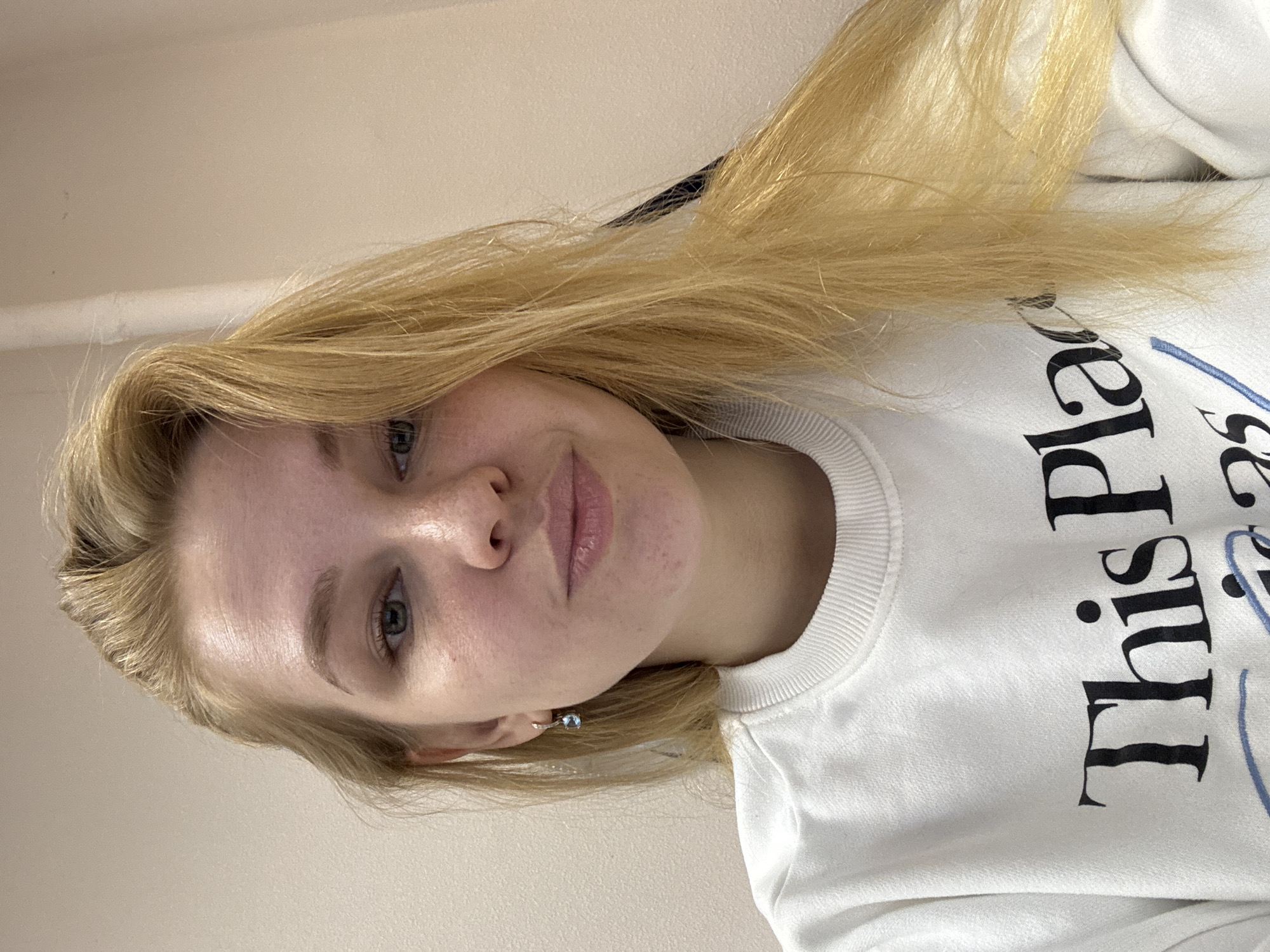 A young woman with blonde hair is posing indoors. She is wearing a white sweatshirt with black lettering that says "This Place" and a light blue design. Her face is visible, showing fair skin, blue eyes, and a slight smile. She is wearing a light blue earring. The background is a plain off-white wall. The photo is taken from a slightly high angle, making the subject look up at the camera. There is no indication of the city Rusanovo or any location in Russia.