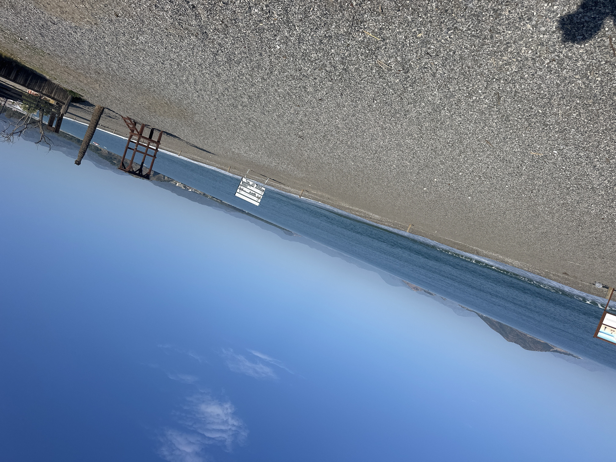 The image depicts a beach in Fethiye, Turkey. The beach is composed of small pebbles. The water of the Aegean Sea meets the shoreline. In the background, mountains are visible along the horizon. A clear blue sky with scattered clouds stretches overhead. There are beach structures along the beach including lifeguard stands. A fence lines the edge of the beach away from the water.
