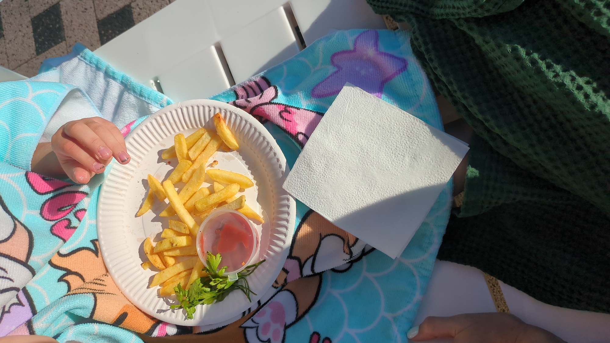 The image shows a plate of french fries and ketchup on a white paper plate, resting on a blue towel with cartoon characters. Next to the plate is a folded white napkin. A child's hand with painted fingernails is visible near the plate, as if reaching for the food. The scene appears to be outdoors, possibly at a park or poolside, indicated by the tiled ground and bright sunlight casting shadows. The towel's design suggests a playful or child-friendly setting. The overall impression is a casual moment of a child enjoying a snack. The location in Vadul lui Voda, Moldova, implies an outdoor recreational area or a private residence with outdoor amenities.