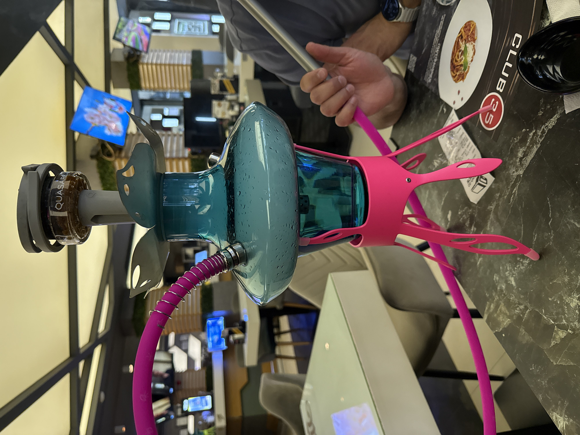 The image depicts a modern hookah setup in what appears to be a lounge or restaurant setting. The hookah features a blue glass base with water droplets, a pink silicone hose, and a pink frame supporting the base. The hookah bowl has the brand "Quasar" visible. A person is holding a metal hookah pipe in their hand, and their arm is visible. On the table, there is a menu with a picture of a pasta dish and the logo "CLUB 35". The surrounding environment features modern architecture with lighting fixtures and video screens displaying images. Considering the logo, it's possible this is a location within Sofia, Bulgaria, assuming the restaurant chain exists there. The table surface appears to be marble or a similar stone material.