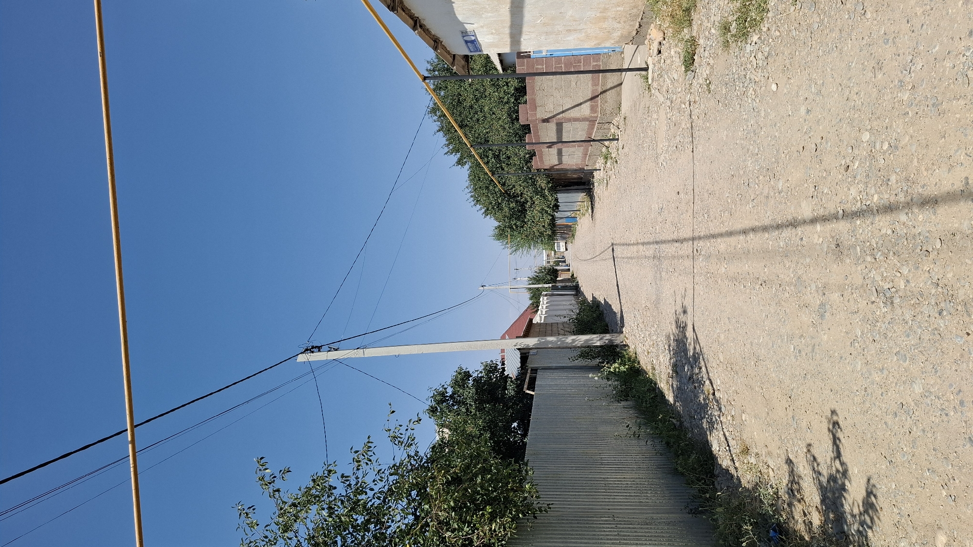 The image shows a dusty, unpaved street in the city of Shubarssu, Kazakhstan, under a clear blue sky. The street is lined with a mix of residential buildings and fences. On the left, a corrugated metal fence runs along the street, with trees and foliage visible behind it. A concrete utility pole with overhead wires stands near the fence. The right side of the street features a brick wall and a building with a weathered exterior. A yellow gas pipe runs horizontally above the street. In the distance, the street curves and appears to continue into a more residential area, with a white van visible. The ground is covered in loose gravel and dirt, with sparse vegetation growing along the sides of the road. The scene suggests a typical day in a provincial town.