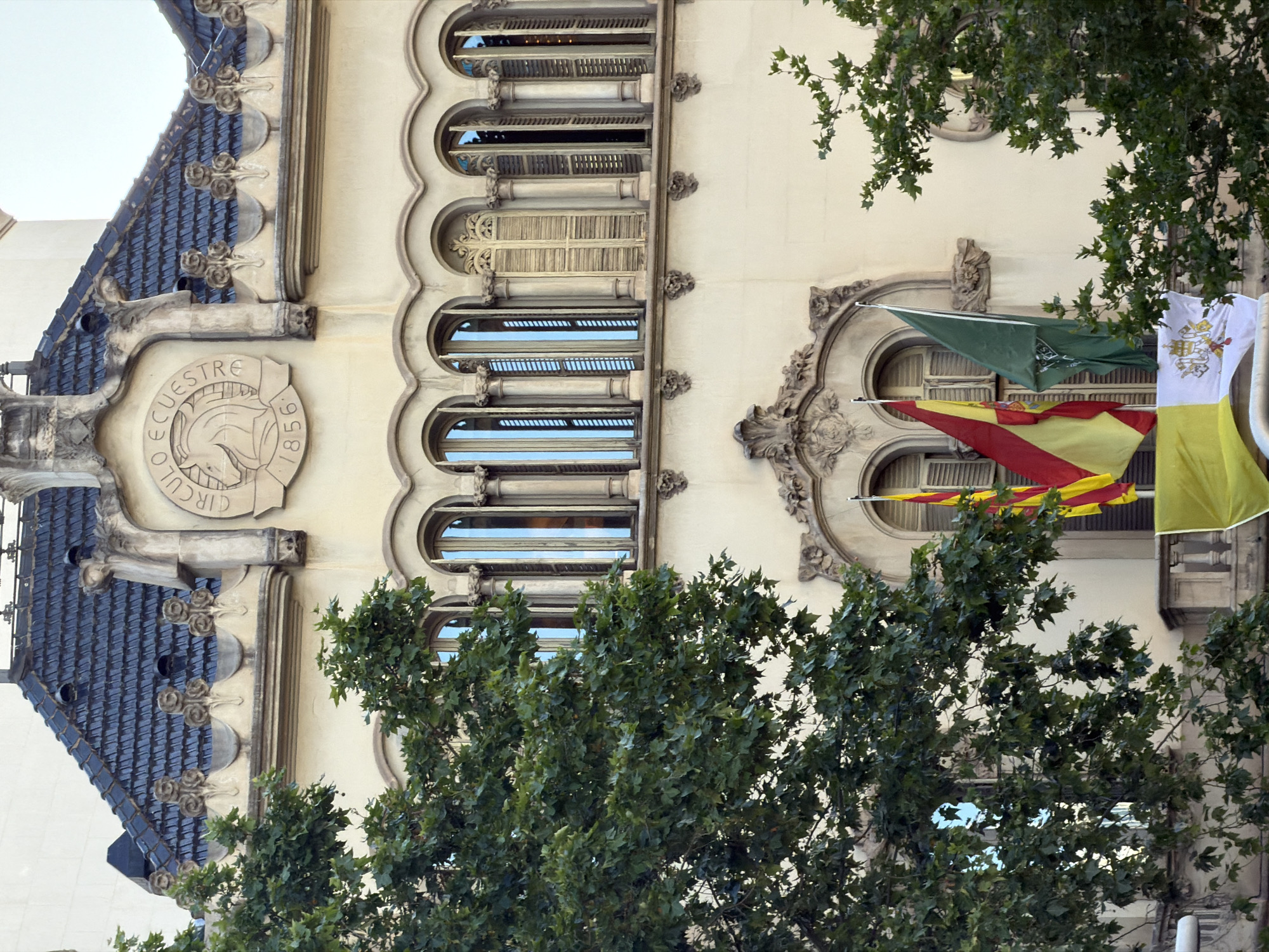 The image displays a detailed view of an ornate building facade in Barcelona, Spain, partially obscured by green tree foliage. The building features a light-colored exterior, likely stone or stucco, with numerous architectural embellishments.
A series of arched windows runs vertically along the facade, some fitted with closed wooden shutters, others reflecting the surroundings. Intricate carved decorations, including floral motifs and scrolled patterns, adorn the spaces between and around the windows. On the upper left, a steeply pitched roof covered in dark blue or grey ceramic tiles is visible, with decorative elements resembling corbels or gargoyles beneath the roofline.
A circular stone crest or medallion is prominently featured on the left side of the facade. It bears a stylized image of a horse's head and is inscribed with the words "CIRCULO ECUESTRE 1856".
On the right, three flags are displayed from an ornate window structure. The uppermost flag is green with some unreadable white text or a logo. Below it is the red and yellow striped flag, identifiable as the Senyera of Catalonia. The bottom flag is yellow and white, featuring a visible coat of arms, possibly representing the Vatican or a papal entity.
Lush green foliage from several trees occupies the foreground and partially covers the building, indicating a daytime setting under clear skies.