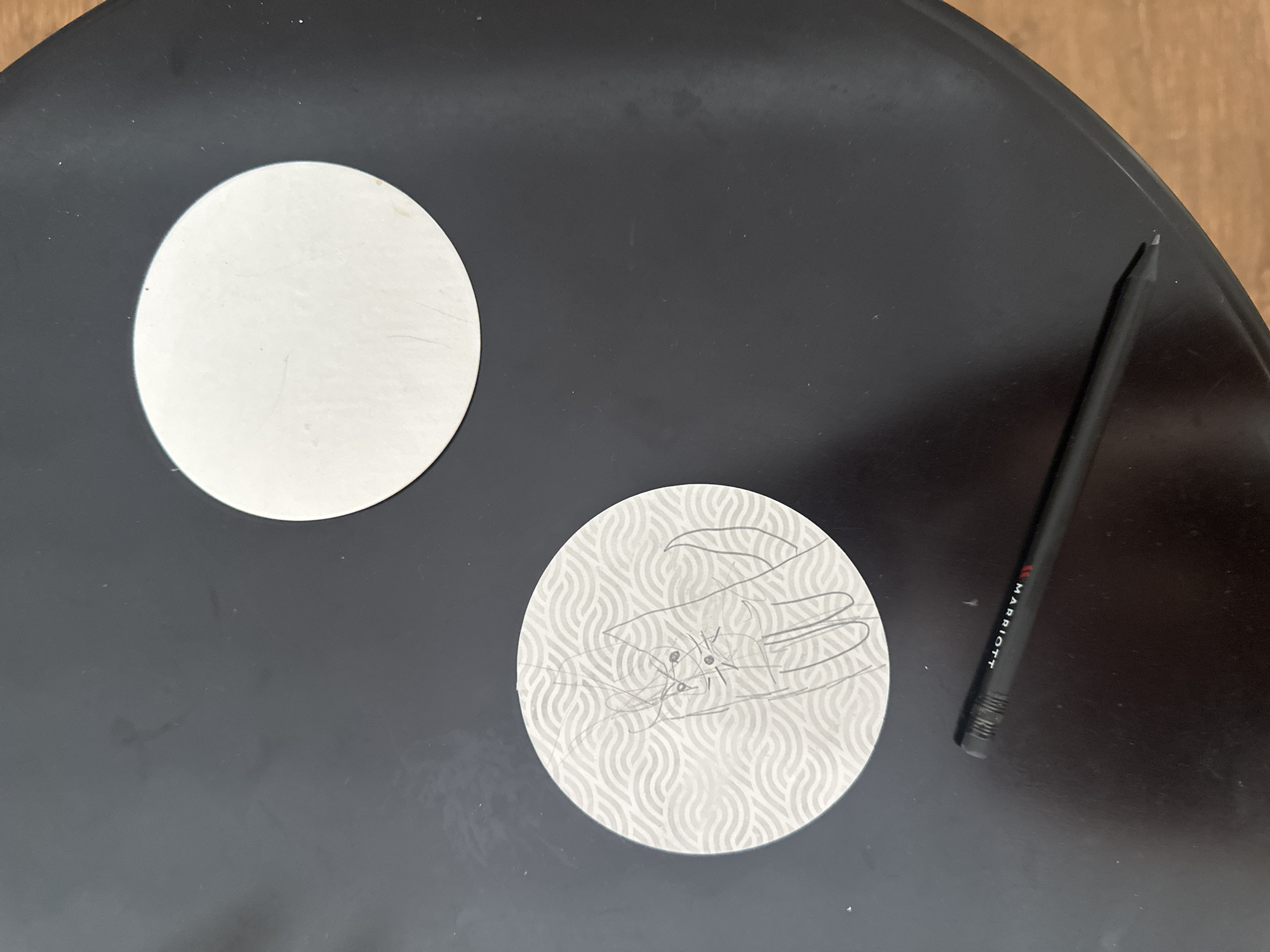The image shows a black, round tabletop. Two coasters are on the table, one plain white and the other with a wavy pattern and a pencil sketch of a cat. A black pencil with "MARRIOTT" printed on it lies next to the patterned coaster. The background is out of focus, but a section of a wooden surface is visible. The location is Unknown, Unknown.