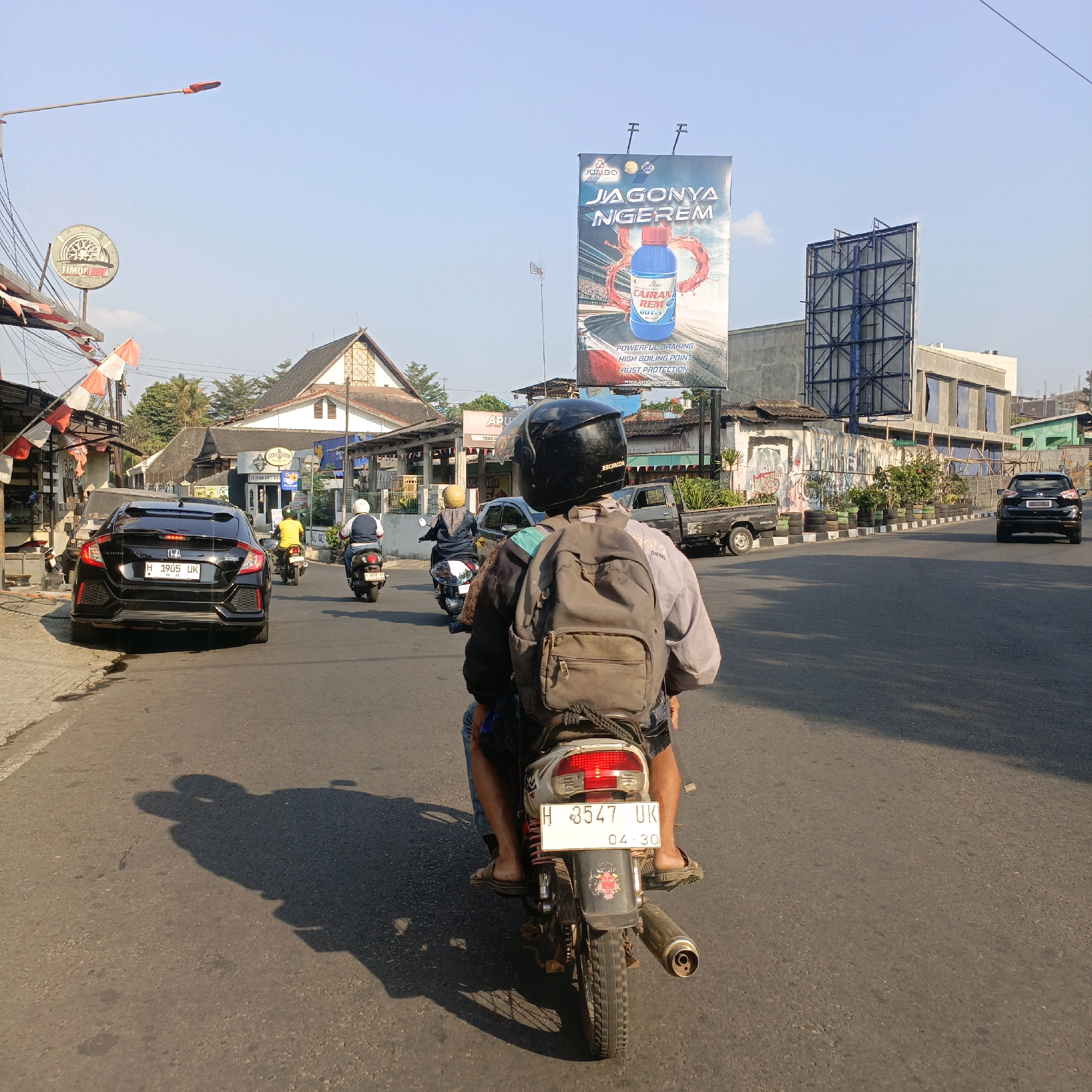The image depicts a street scene in Salatiga, Indonesia, on a clear day with bright sunlight casting long shadows. In the foreground, two people are riding a motorcycle, viewed from behind. The rider wears a black helmet, and a large grey backpack is strapped to the rear passenger. The motorcycle has a white license plate with the text "H 3547 UK" and "04.30". The rear passenger is wearing a light-colored jacket and shorts. They are traveling on a paved road alongside other vehicles, including a black Honda Civic parked on the left with its license plate reading "H 1905 UK". Further down the road, two other motorcycles are visible, carrying riders with helmets. To the right, a black pickup truck is parked.
The surrounding environment includes buildings lining the street, some with traditional Indonesian architecture, and a large billboard in the distance advertising "JAGONYA NGEREM" with an image of a bottle of brake fluid. Power lines and a street lamp are also visible. The overall impression is of a typical urban street scene during daylight hours.