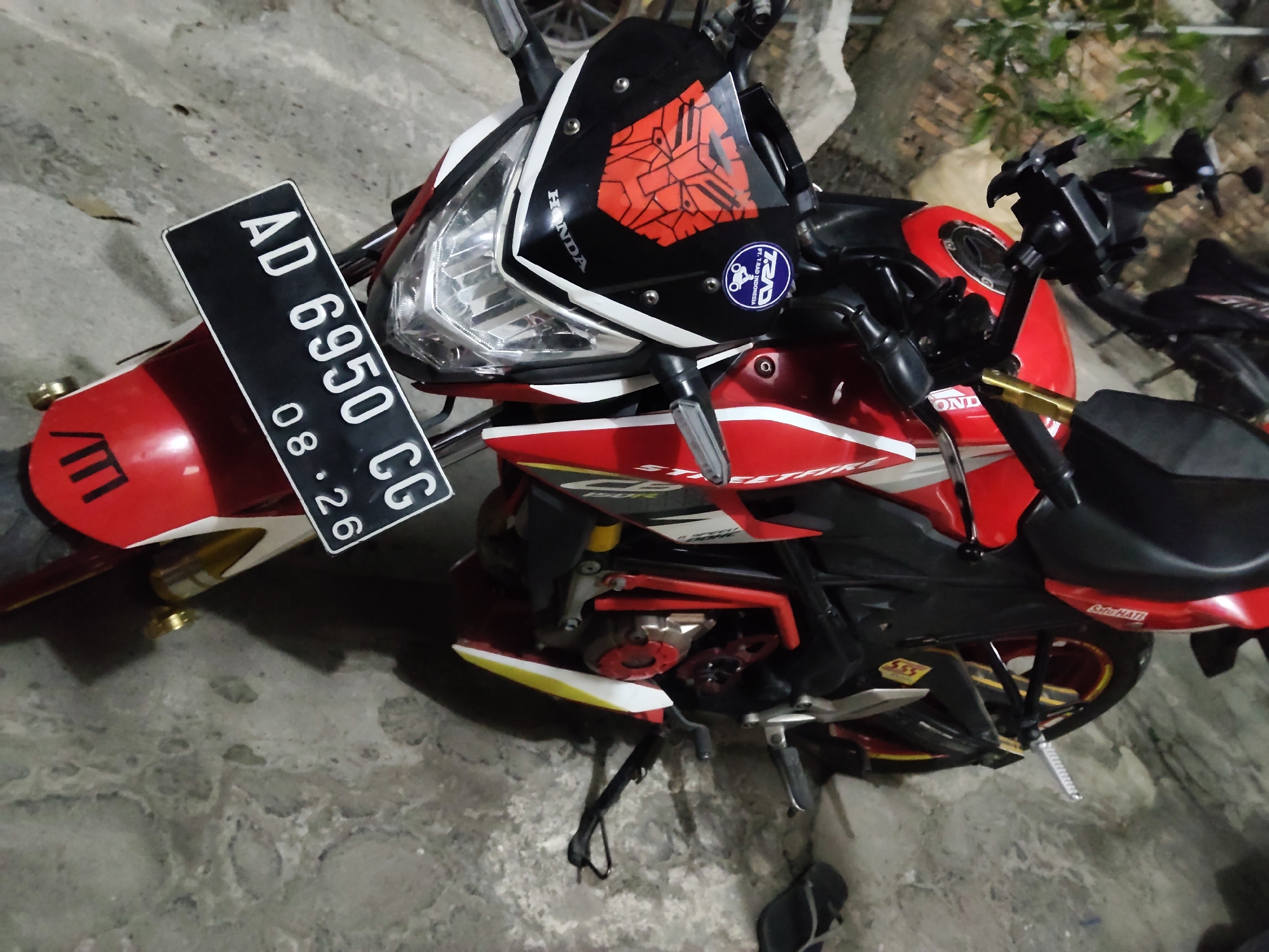 A red and white Honda motorcycle is parked on a concrete surface. The motorcycle has a black license plate that reads "AD 6950 CG 08.26". There is a sticker on the front fairing that appears to be a red geometric design. The motorcycle is outdoors, with some foliage visible in the background. The scene is located in Mekarmukti, Indonesia.