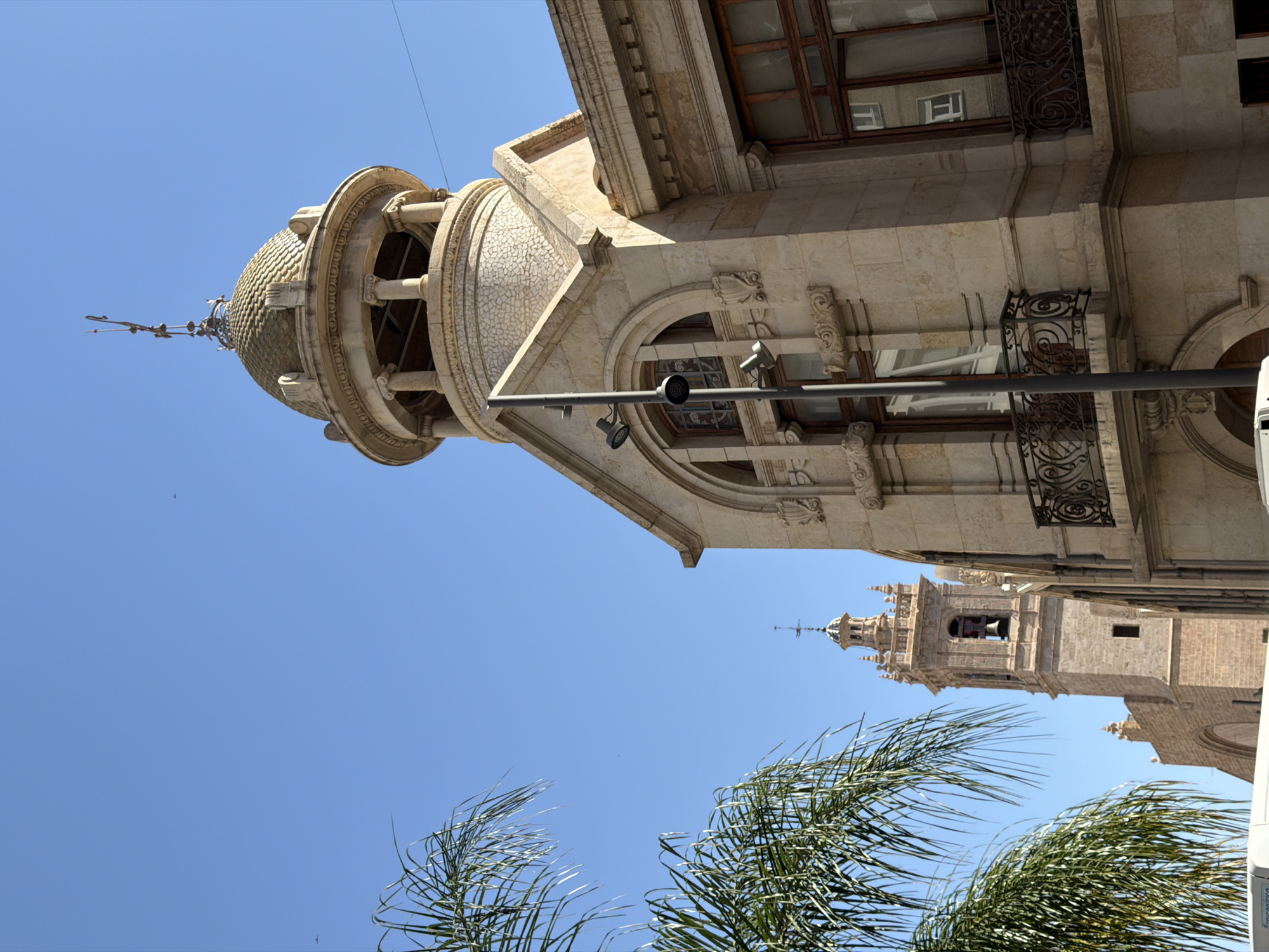 A low-angle, medium shot captures a pale blue sky and the upper portions of two ornate buildings in an urban environment. On the right, a light-colored stone building features a domed roof topped with a decorative finial, framed by stone columns and arches. To the left, another building with a tall bell tower, constructed from a similar light stone, rises against the sky. Palm fronds from a tree are visible in the foreground on the left, partially obscuring the view. The scene appears to be a sunny day with good visibility.